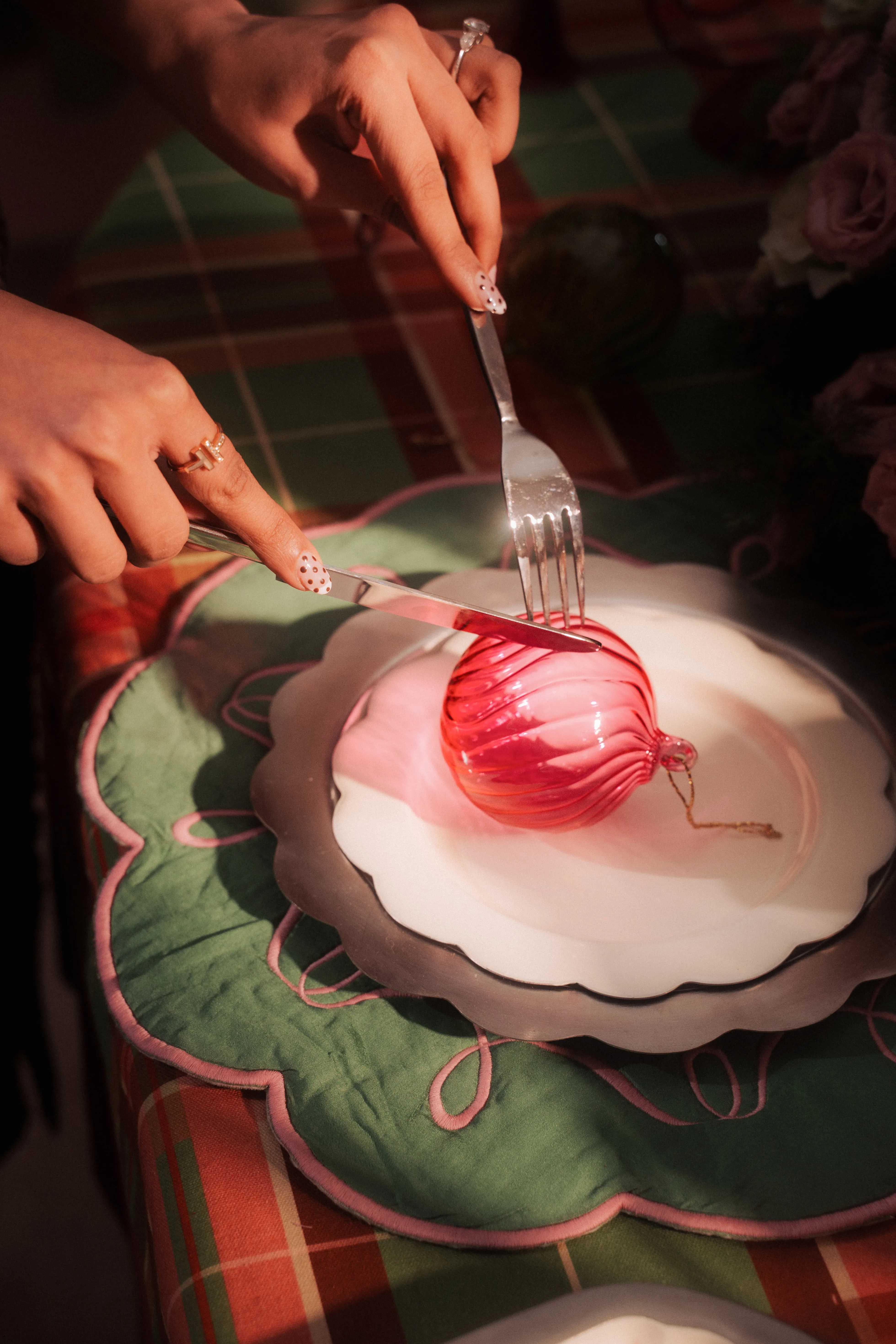 Hands using a knife and fork to cut into a magenta glass ball ornament plated like a dessert on a scalloped plate.