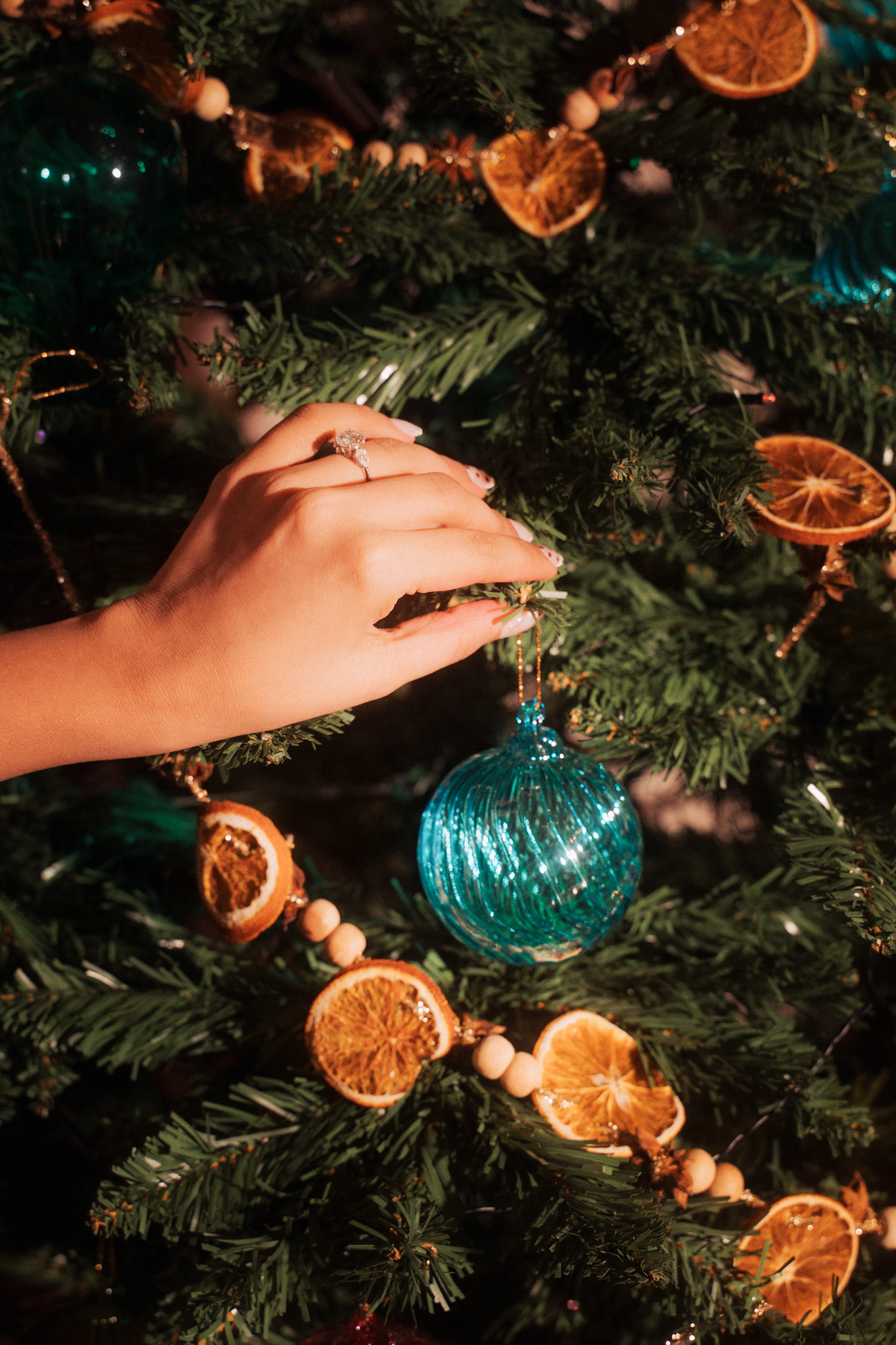 A hand reaching to hang a teal glass ball ornament on a Christmas tree strung with dried orange slices.