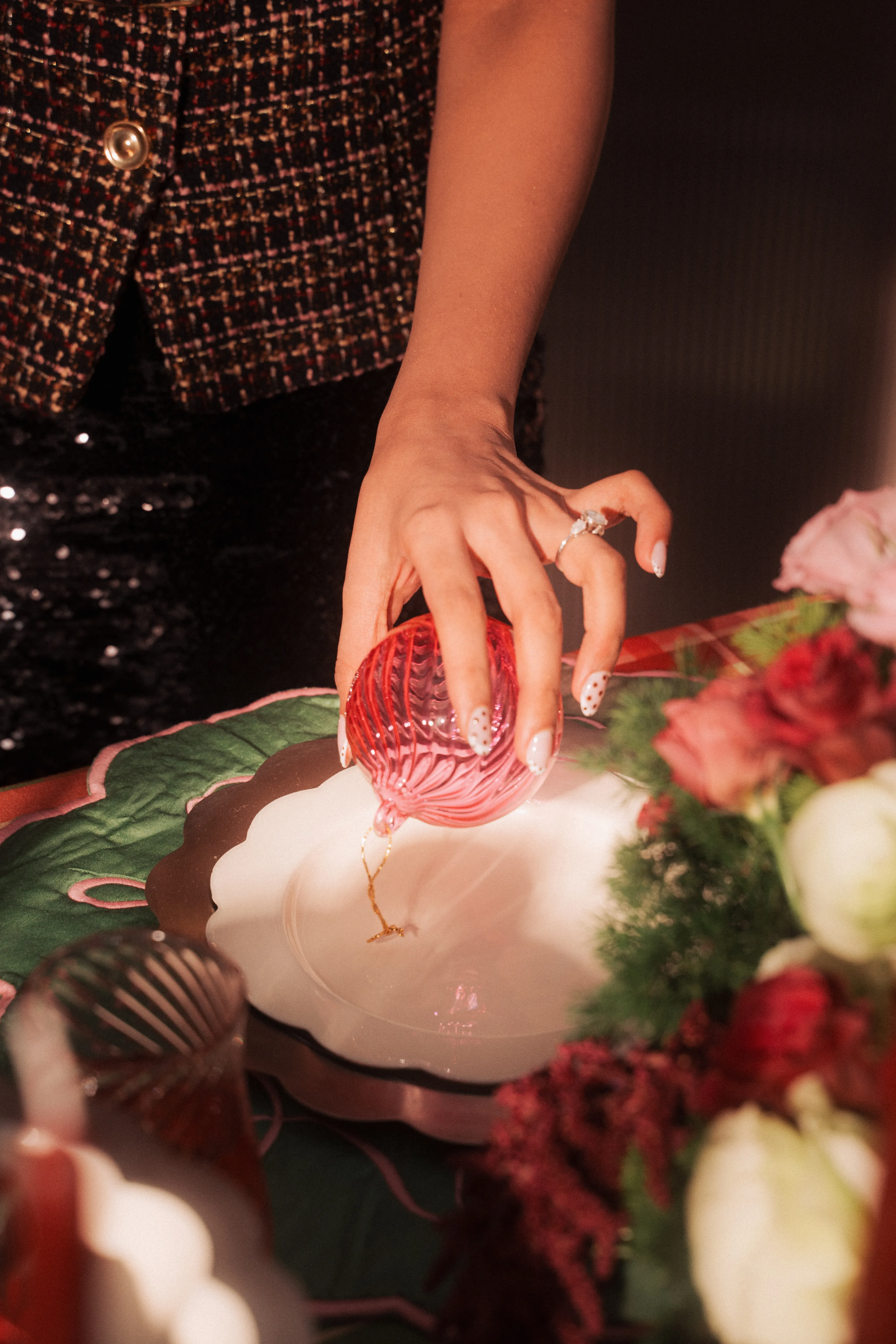 A hand in a sequined sleeve setting a red glass ball ornament onto a plate beside a floral arrangement.