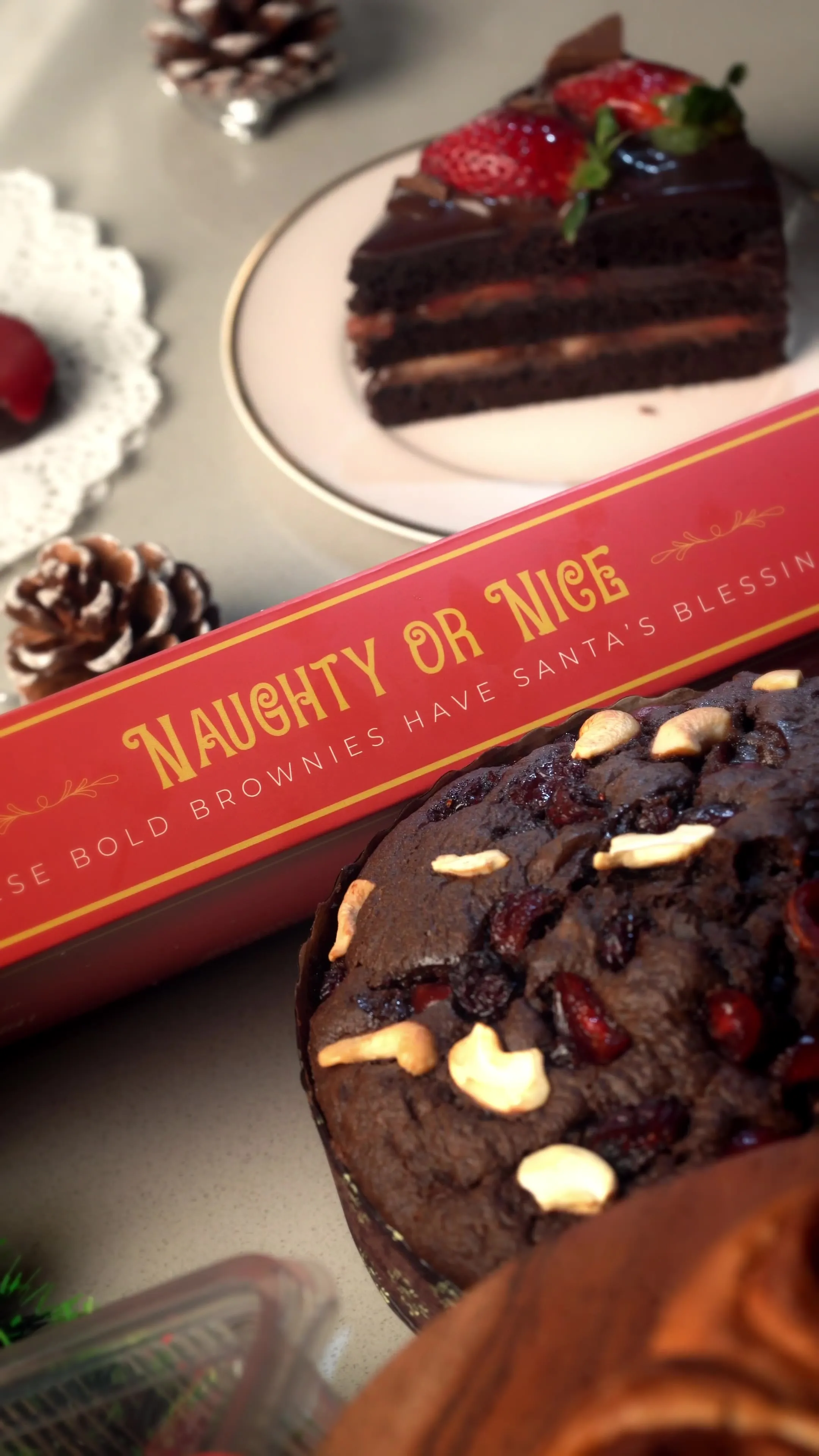 A slice of layered chocolate cake topped with strawberries beside a red "Naughty or Nice" Bold Brownie Lab box and a cashew-topped fruit cake.