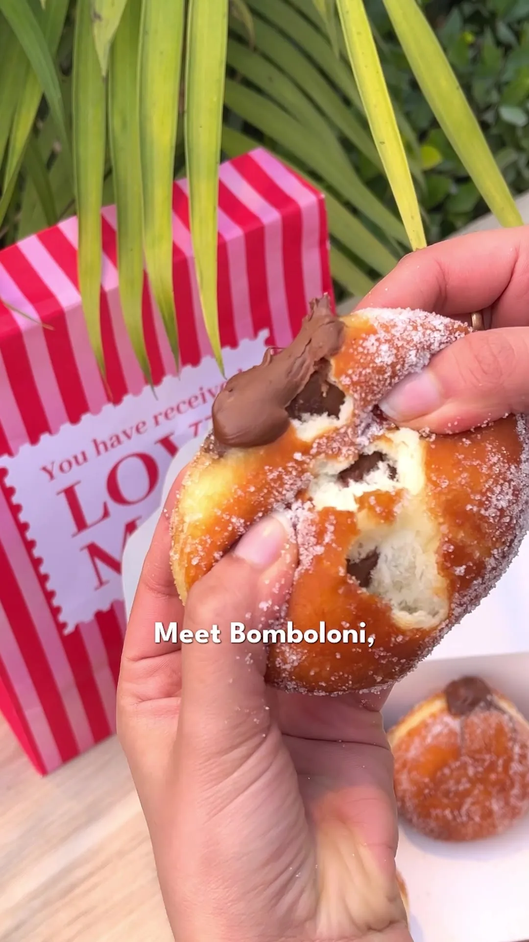 A hand holding a torn-open, Nutella-filled bomboloni beside a striped gift bag, captioned "Meet Bomboloni."