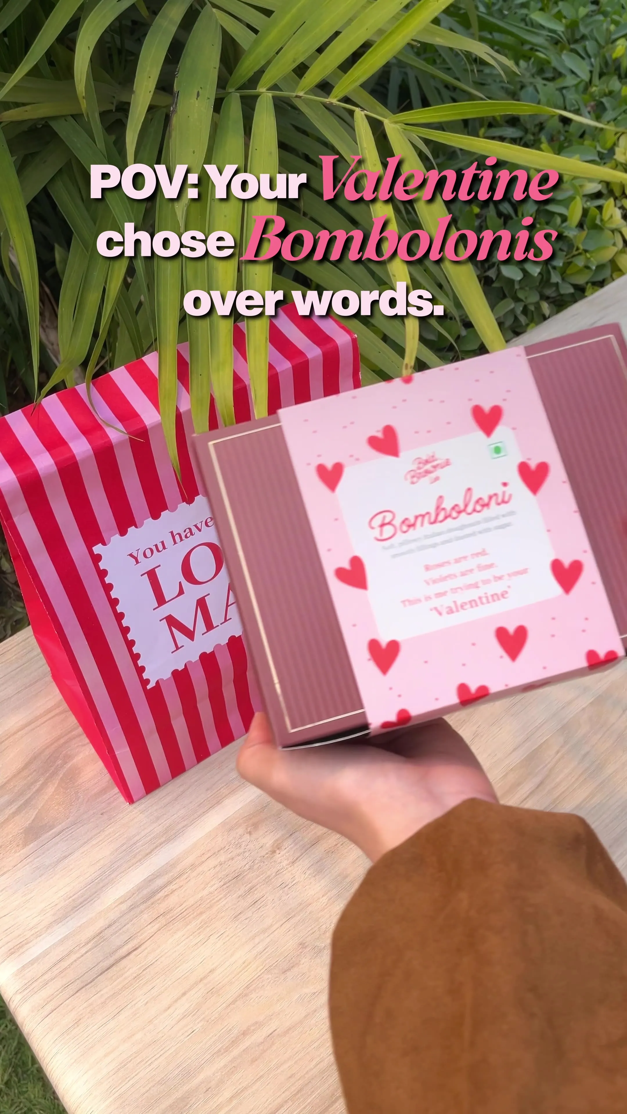 A heart-patterned Bomboloni gift box held up in front of a striped bag, captioned "POV: Your Valentine chose Bombolonis over words."