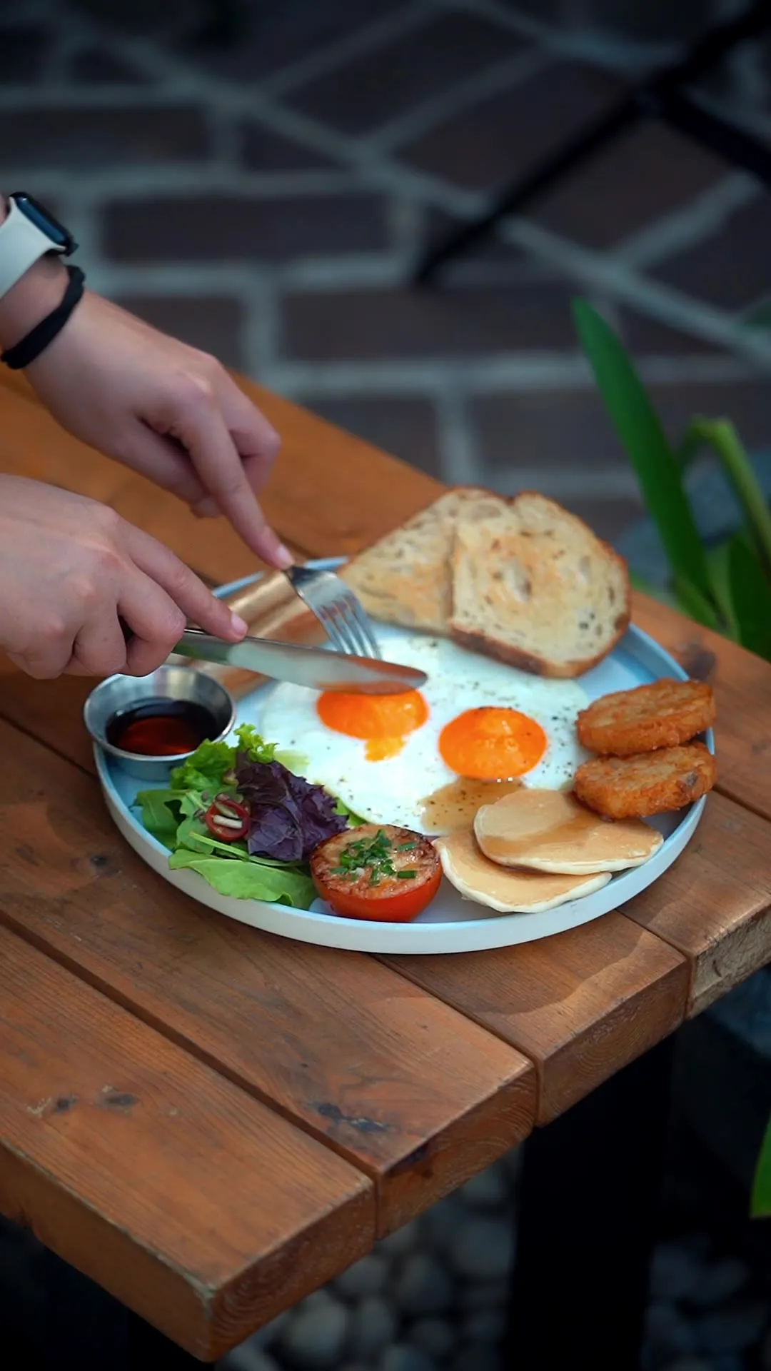 Hands cutting into a fried egg breakfast plate with toast, sausages, and salad.