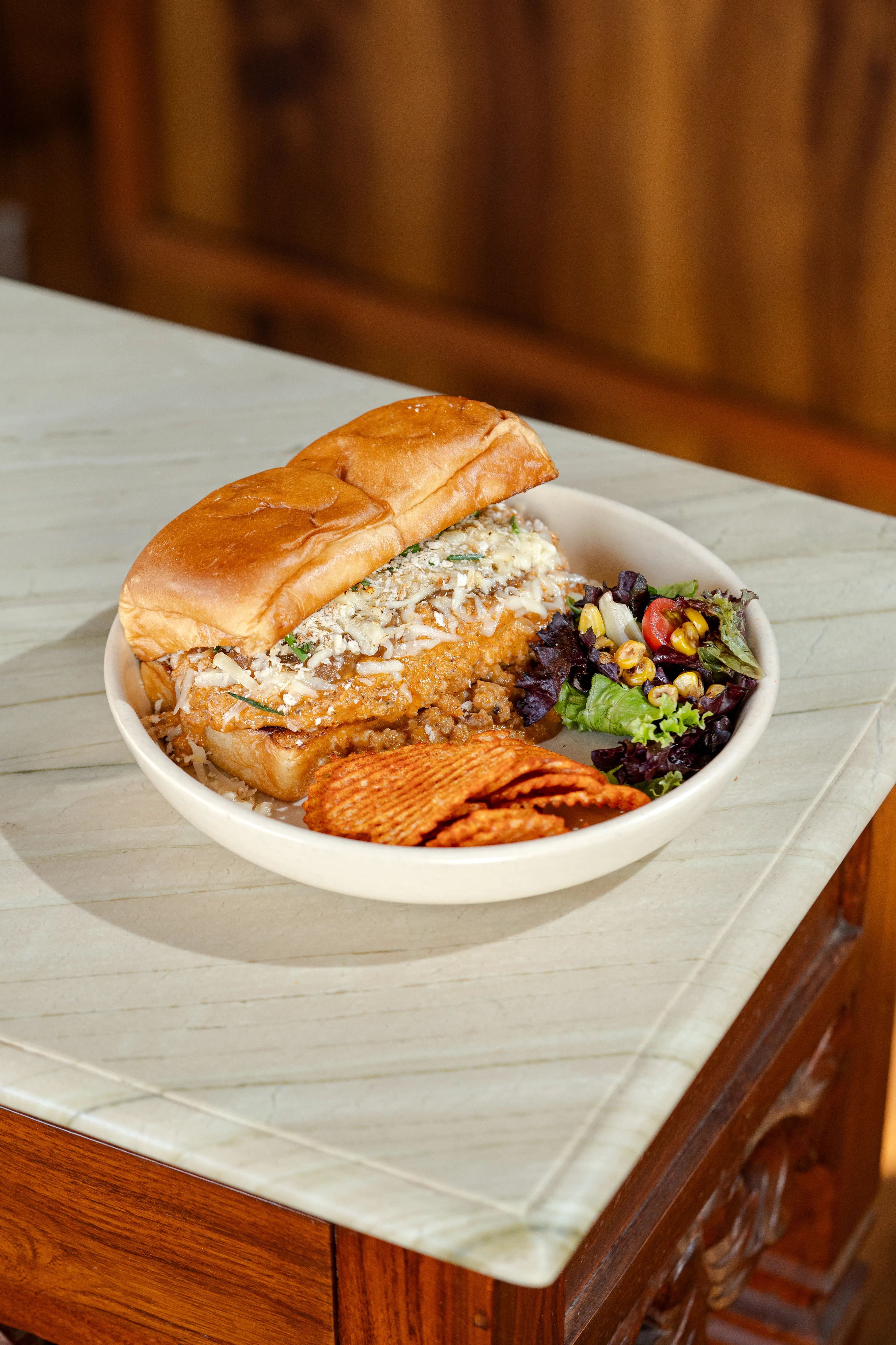 A toasted bun sandwich topped with melted cheese, served in a bowl with a corn-and-tomato salad and spiced ridged chips.
