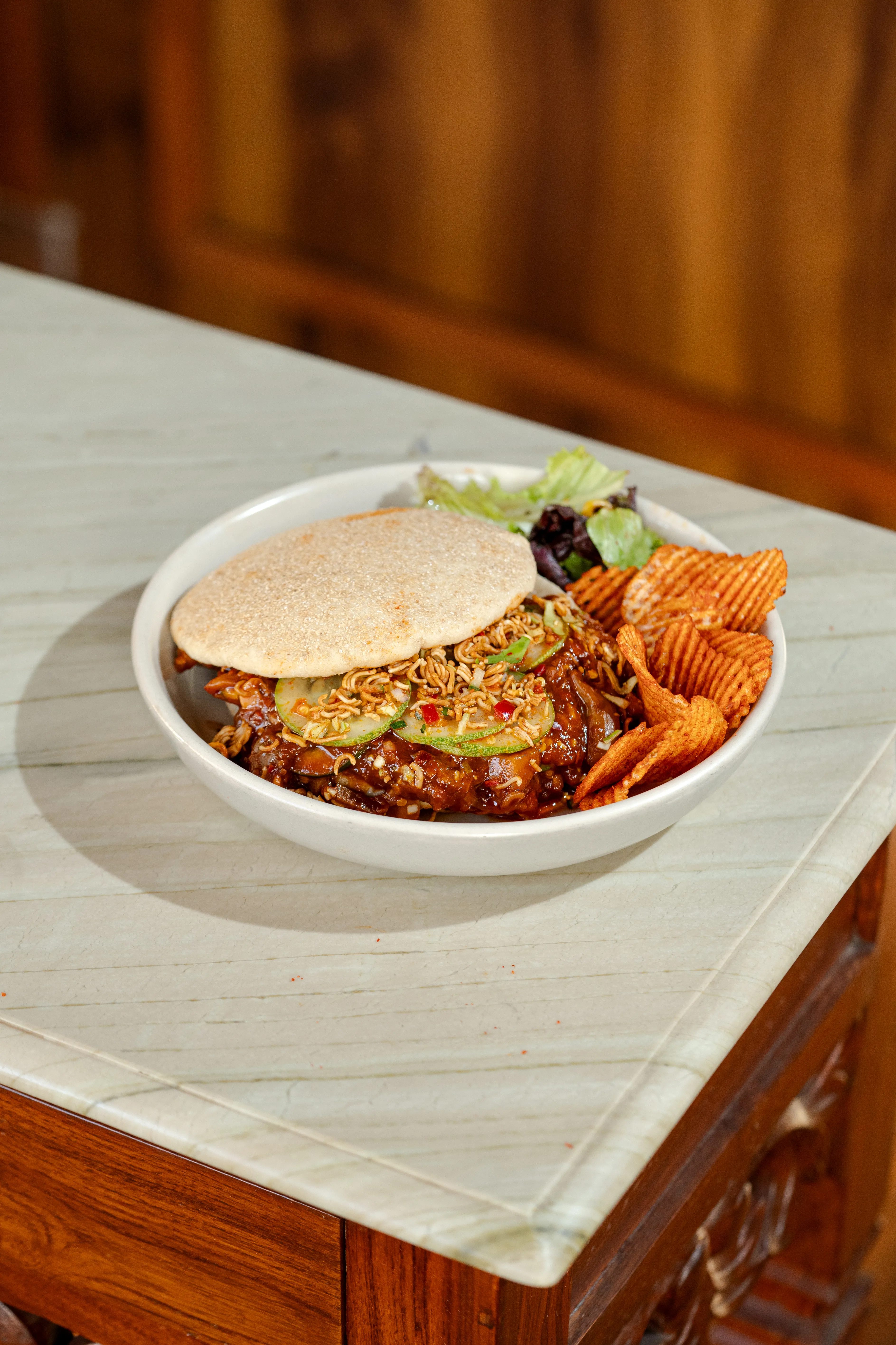 A flatbread-topped sandwich piled with ramen noodles and cucumber slices in chili sauce, served with salad and chips.