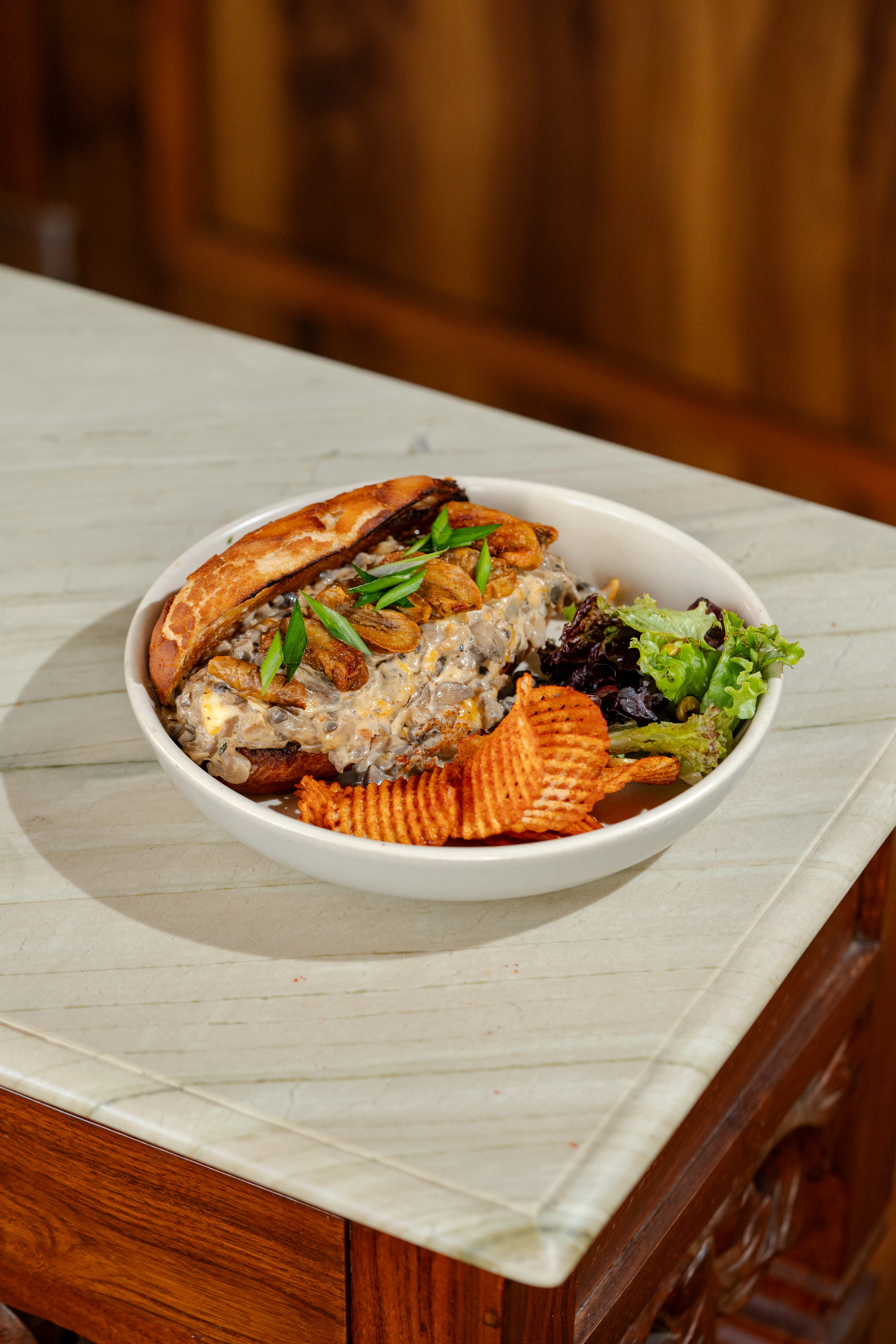 A mushroom and chicken melt sandwich with scallion garnish in a bowl, served with mixed greens and spiced chips.