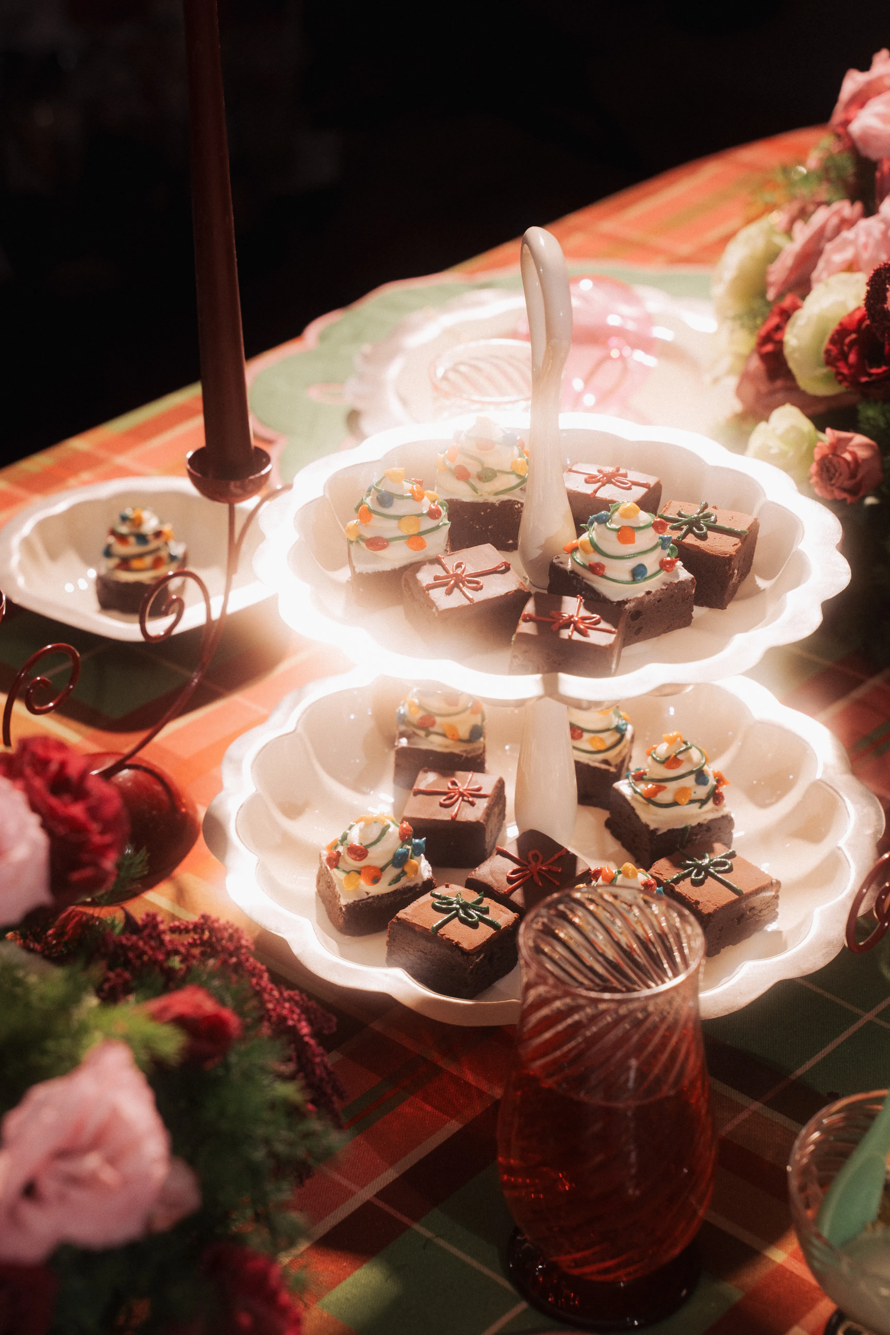 A tiered white cake stand of gift- and tree-decorated brownie bites beside a candle holder and a glass of iced tea on a plaid tablecloth.