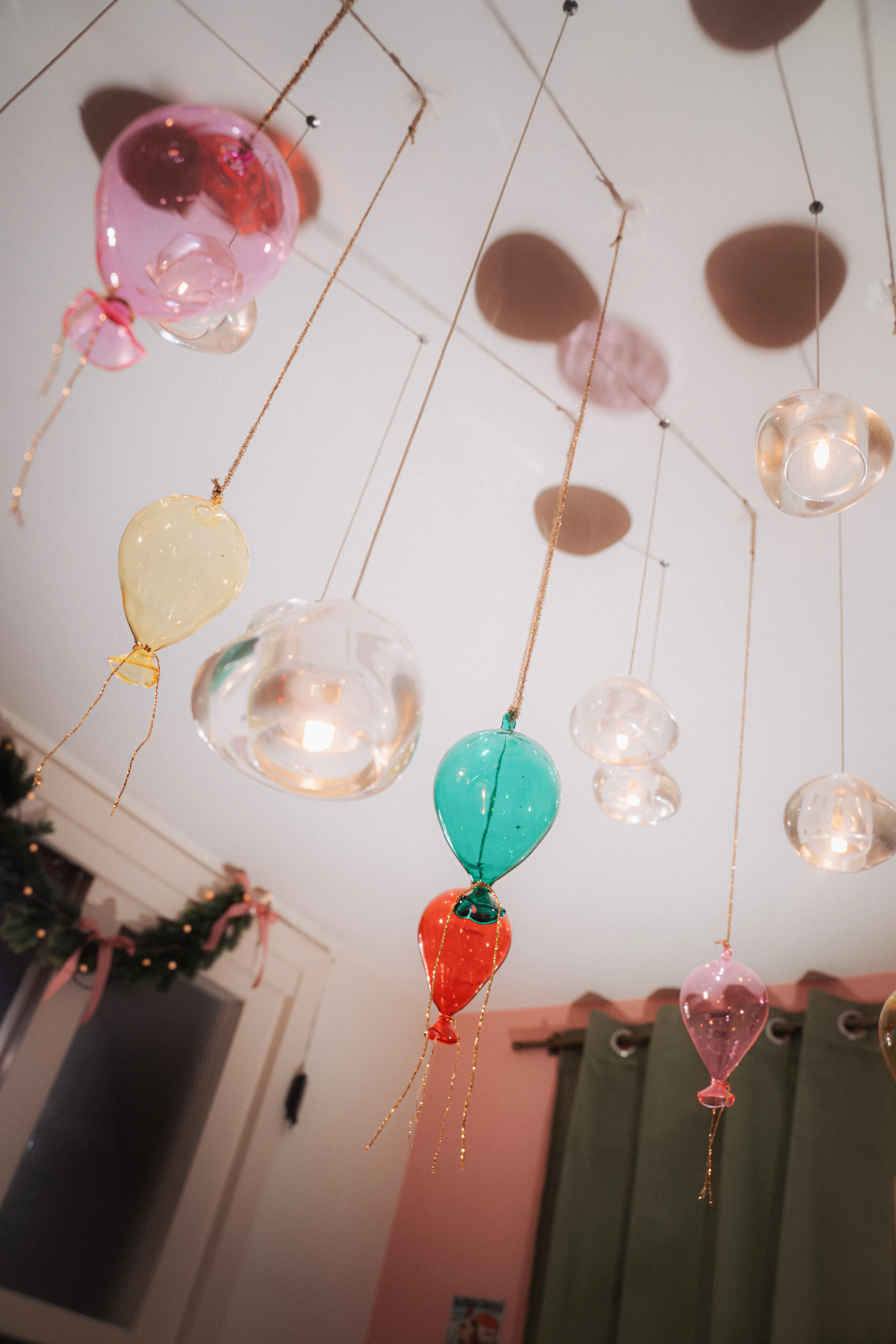 Glass balloon- and bubble-shaped ornaments in pink, yellow, teal, and clear hanging from strings against a white ceiling.
