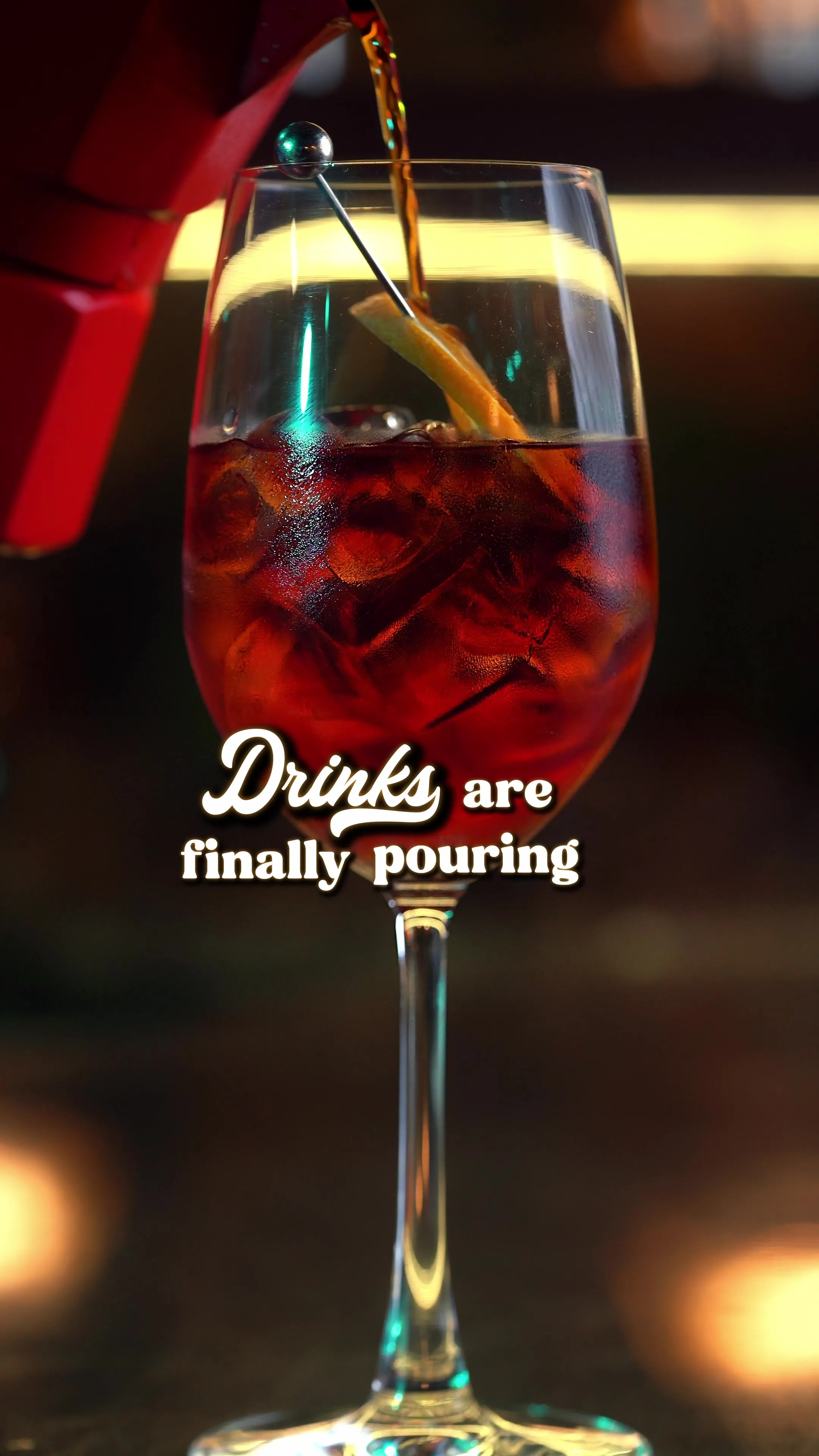 A red cocktail being poured into a glass with an orange garnish, captioned "Drinks are finally pouring."