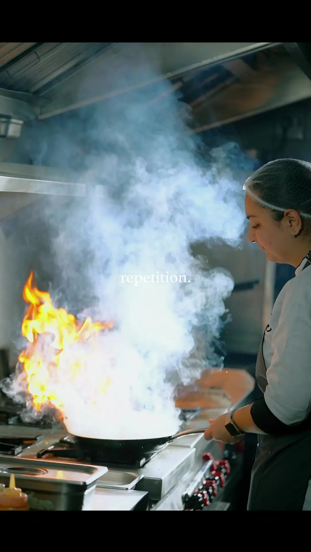 A chef working in the kitchen amid rising flame and smoke, captioned "repetition."