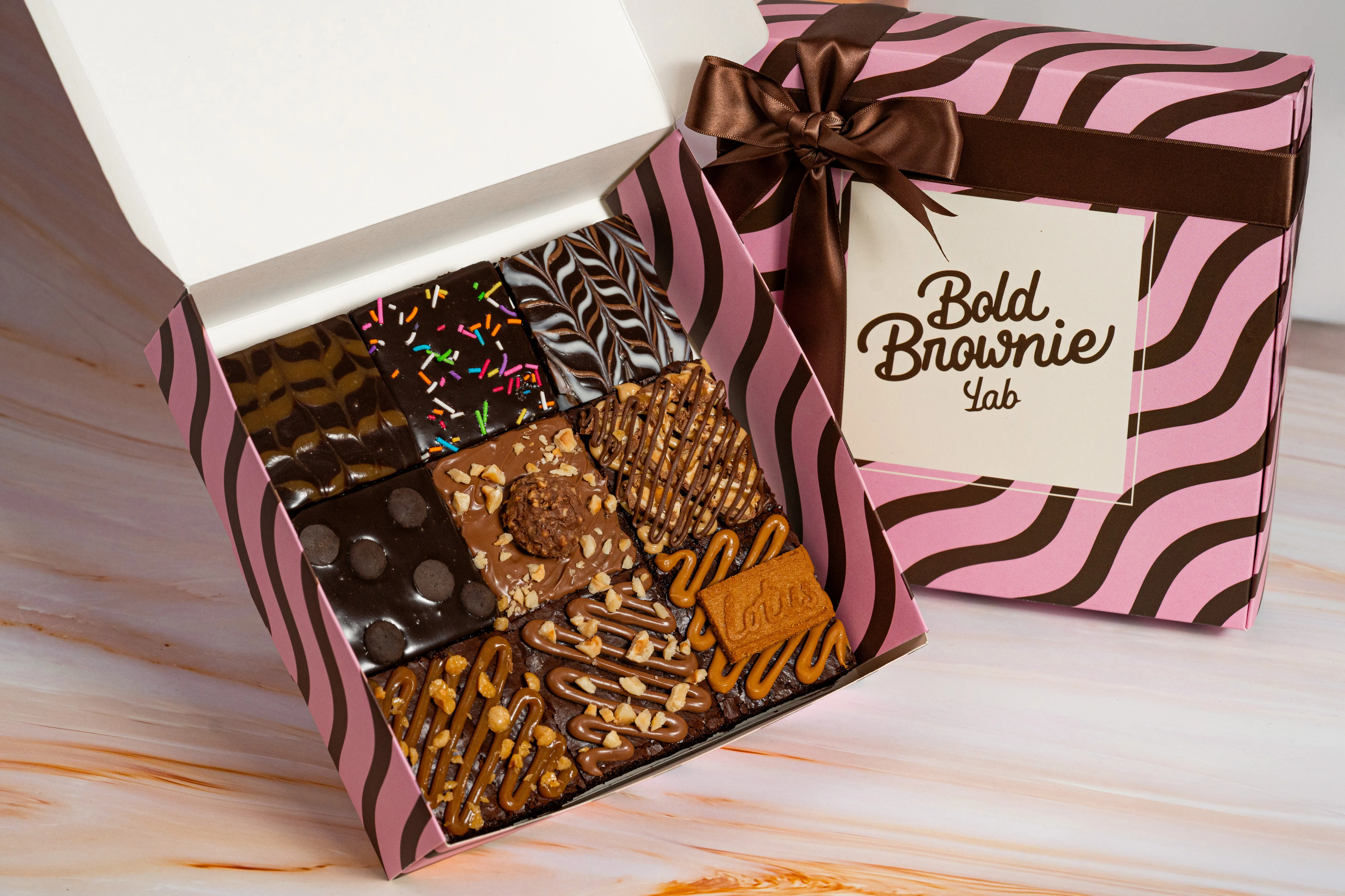 An open Bold Brownie Lab box showing six assorted brownies — a chocolate swirl, rainbow sprinkles, a hazelnut-and-truffle top, and a peanut butter-and-Lotus-biscuit top — beside the ribboned box lid.