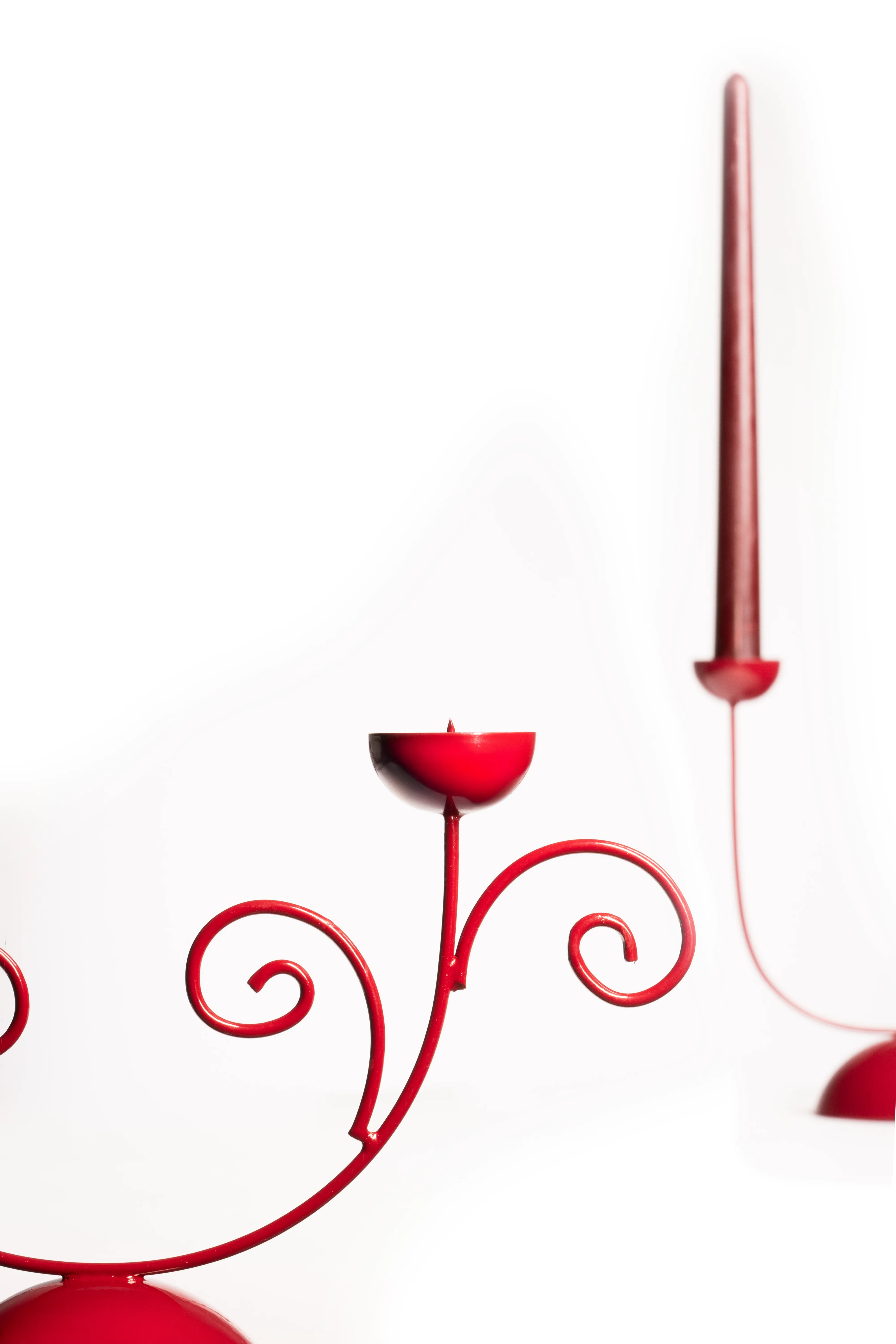 A red scrollwork double candle holder shot at an angle, its dome base and a taper candle visible against a white background.