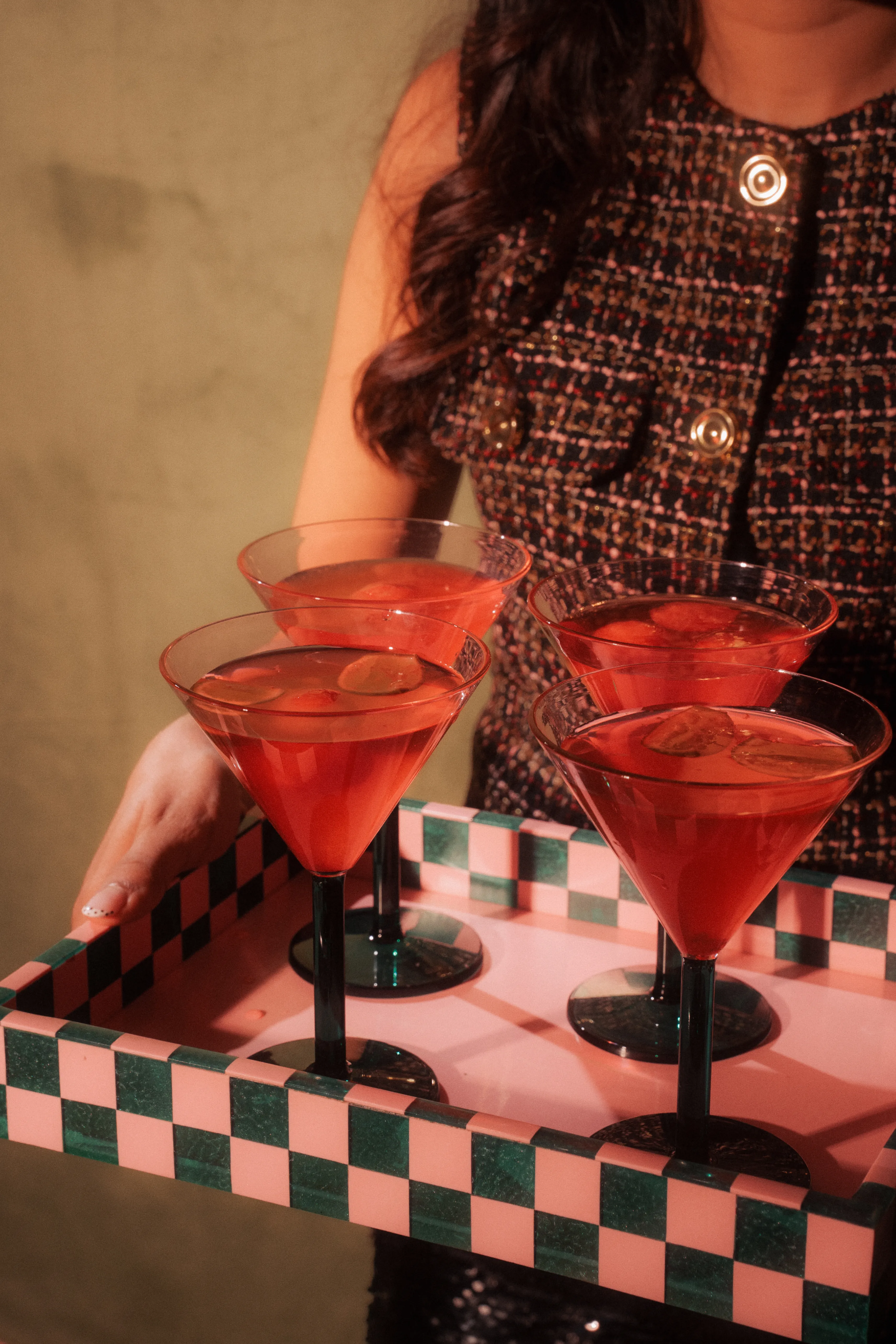 A woman in a tweed dress holding a checkered tray of four red cocktails in coupe glasses.