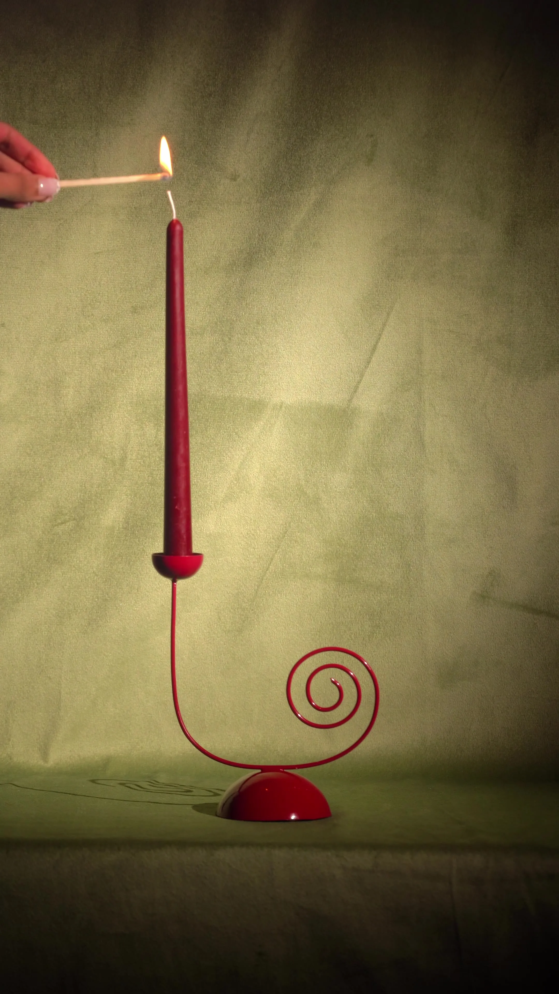 A hand lighting a red taper candle set in a red spiral candle holder against a green backdrop.