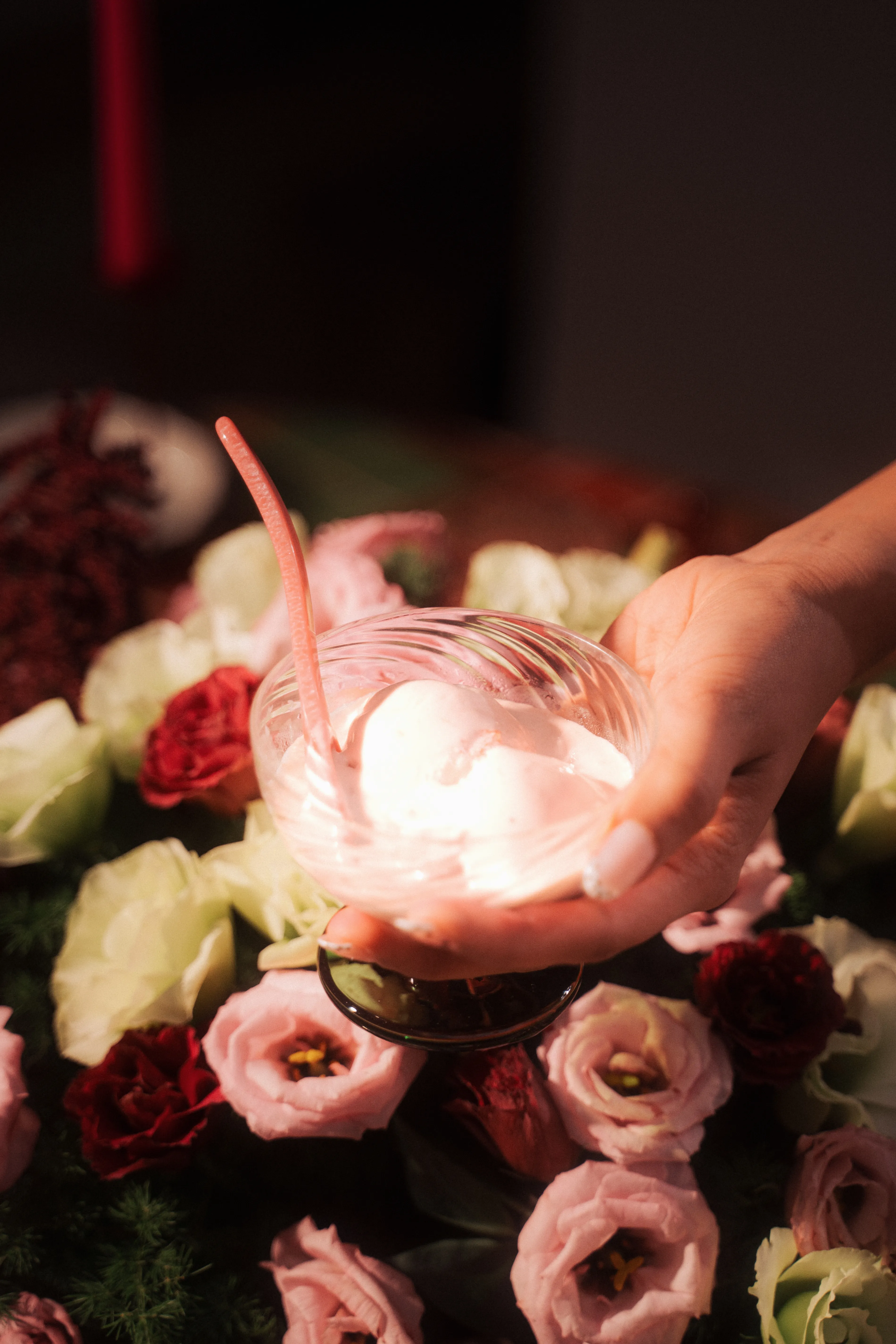 A hand holding a ribbed glass cup of whipped dessert with a pink spoon, resting on a bed of pink and white roses.