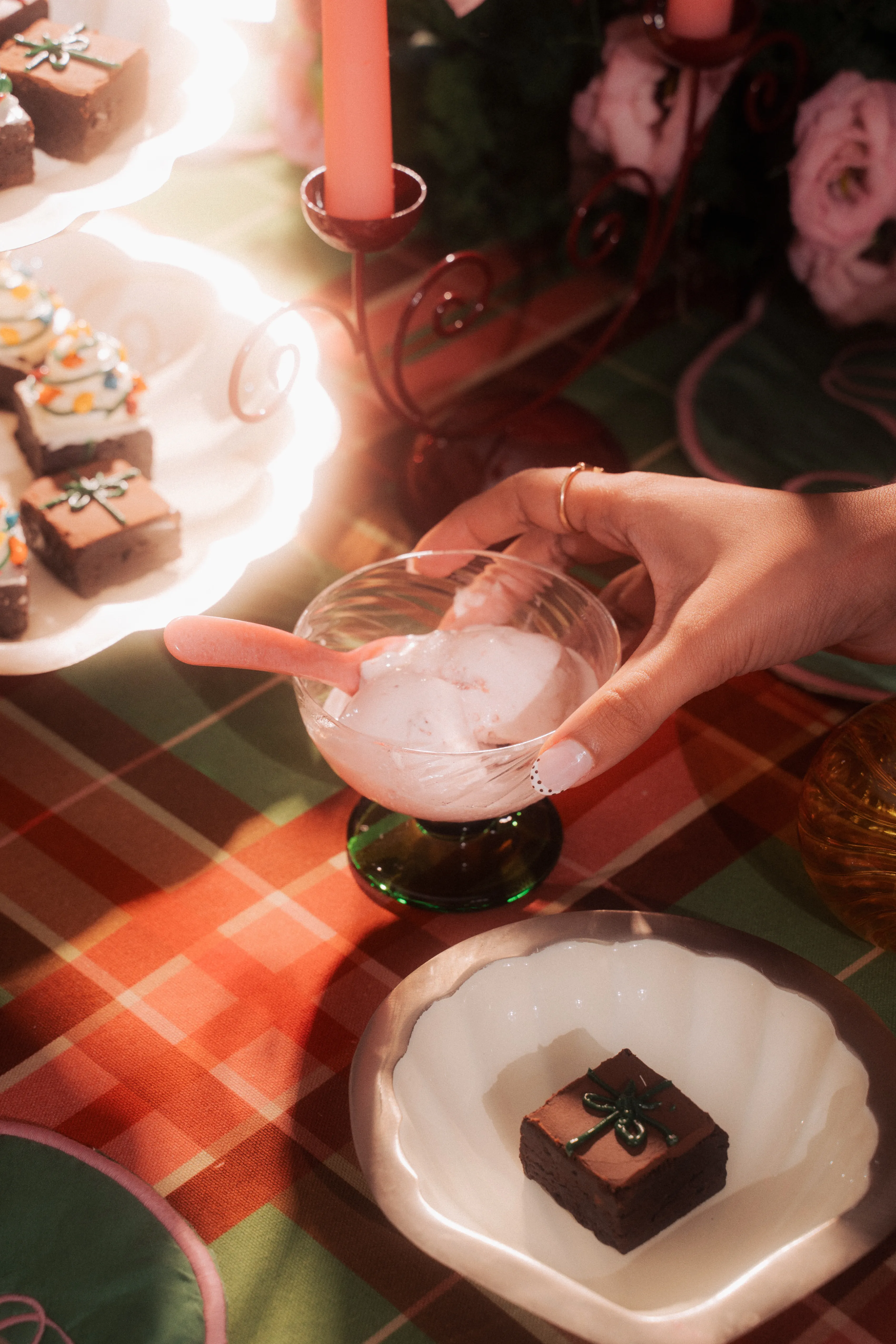 A hand holding a glass of whipped dessert beside plates of tree-decorated brownie bites and a lit red candle holder.