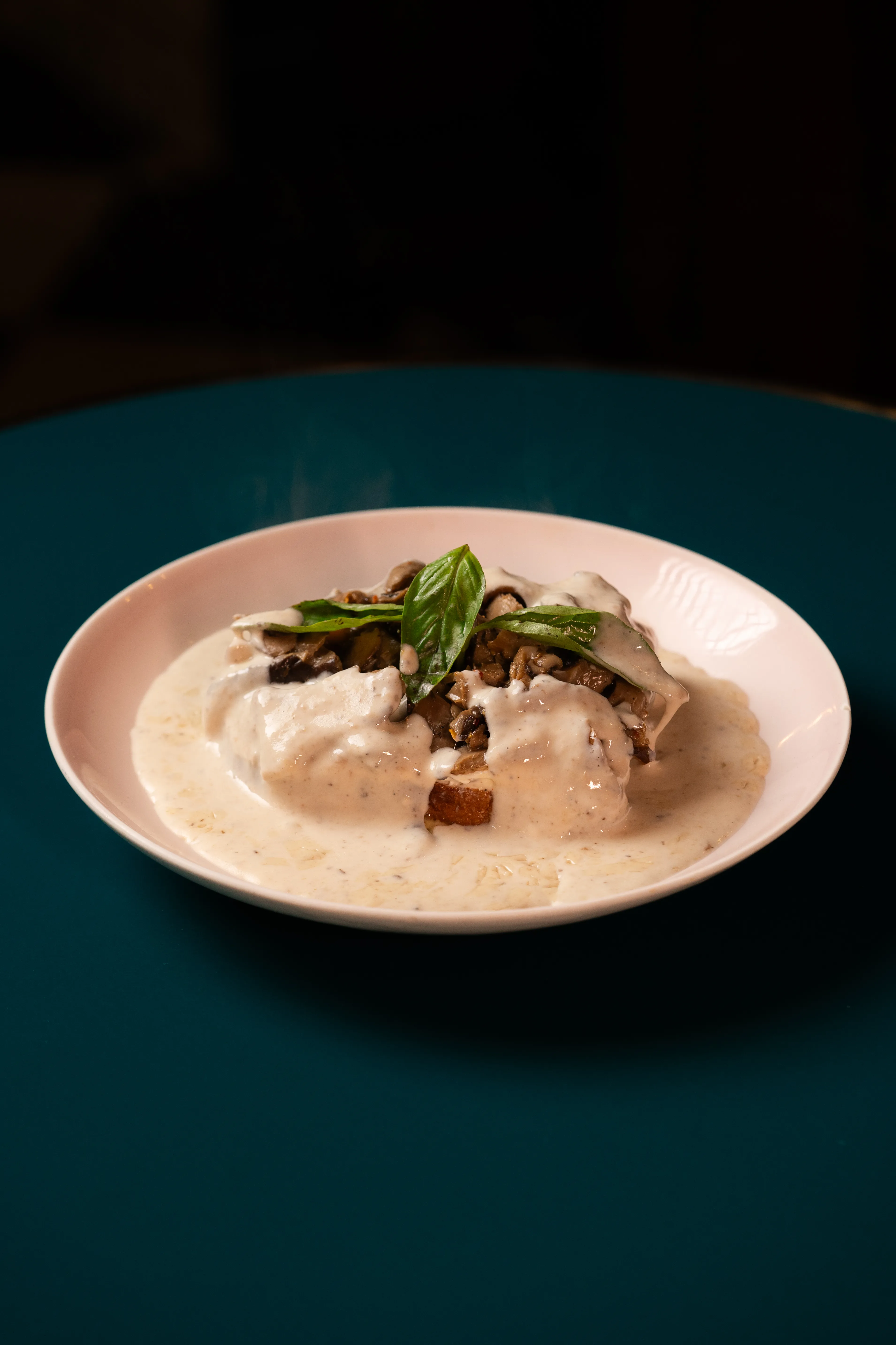 A creamy mushroom toast dish garnished with basil, plated on a pink dish against a dark teal table.