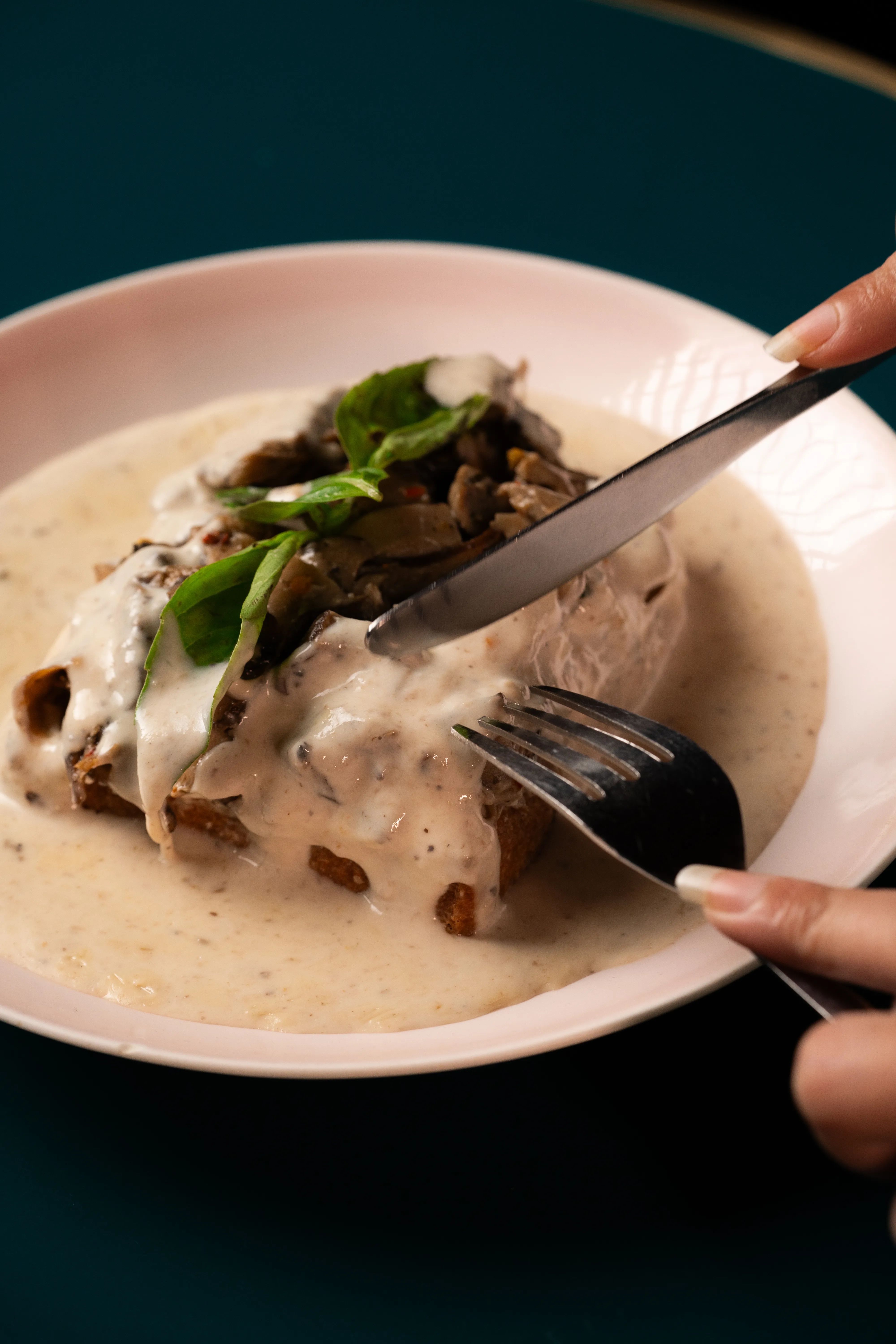 A knife and fork cutting into the creamy mushroom toast, basil garnish visible.