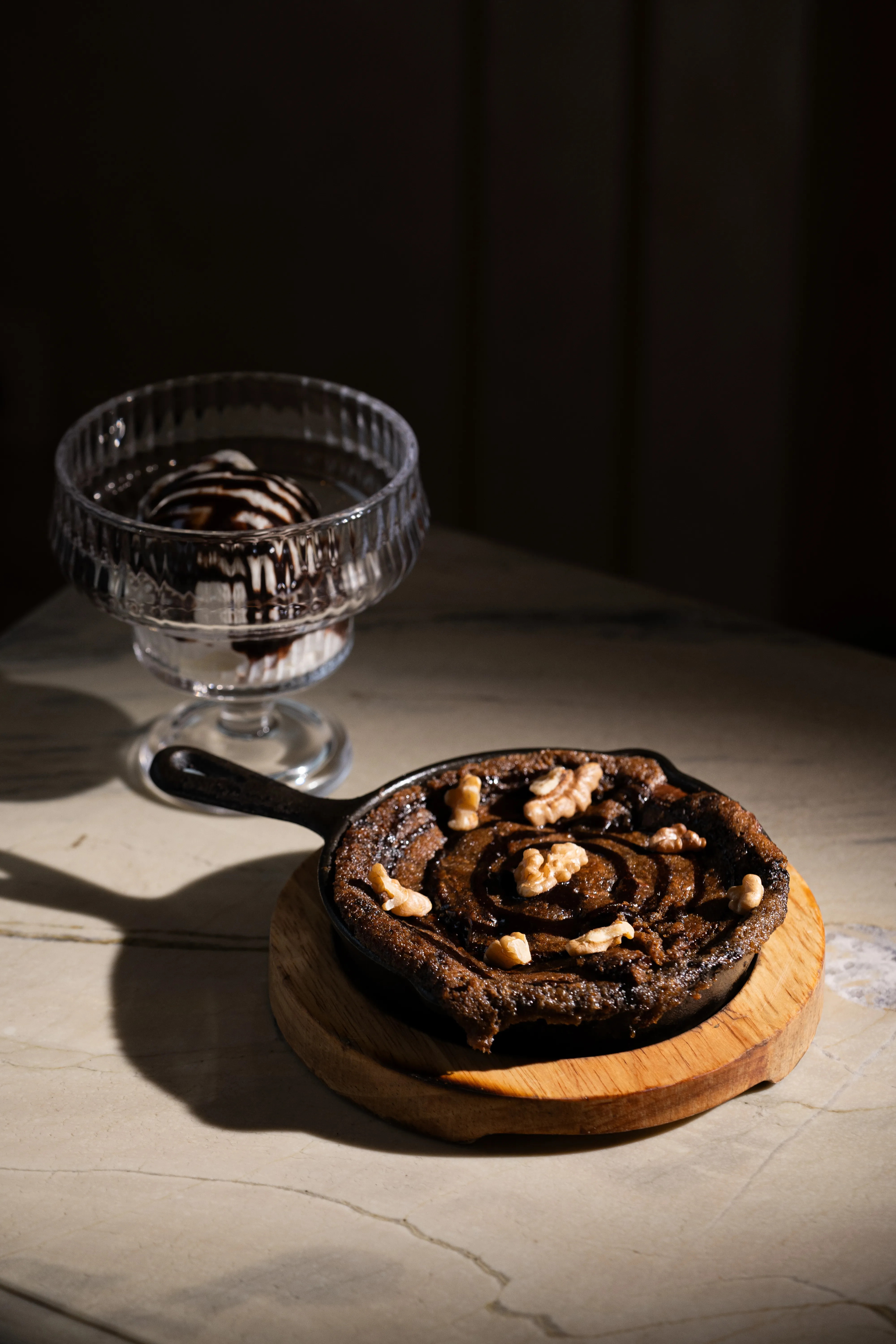 A walnut skillet dessert beside a glass sundae cup of ice cream drizzled with chocolate, on a sunlit marble table.