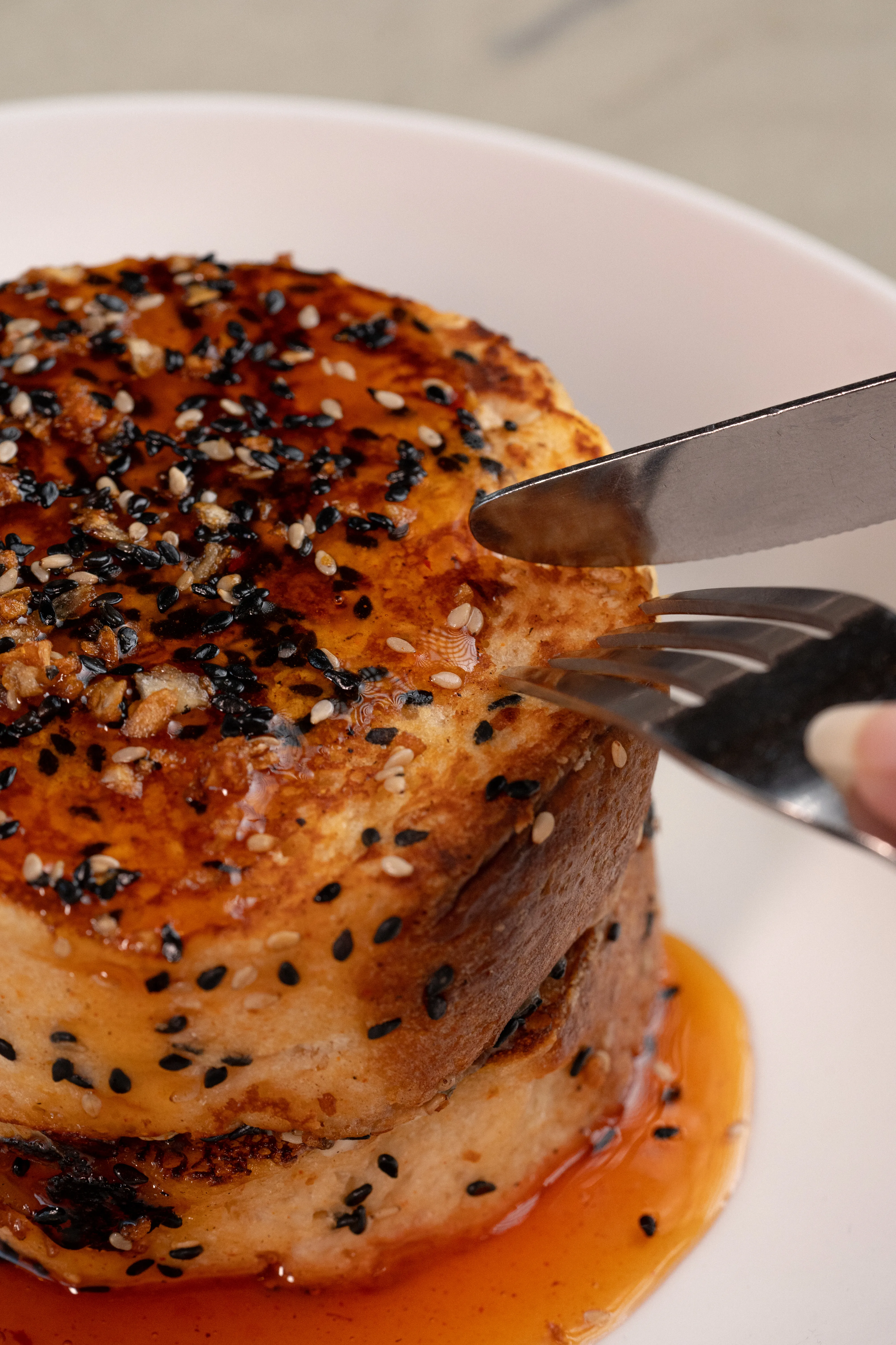 A stack of black sesame pancakes glazed in caramel sauce, being cut into with a fork and knife.