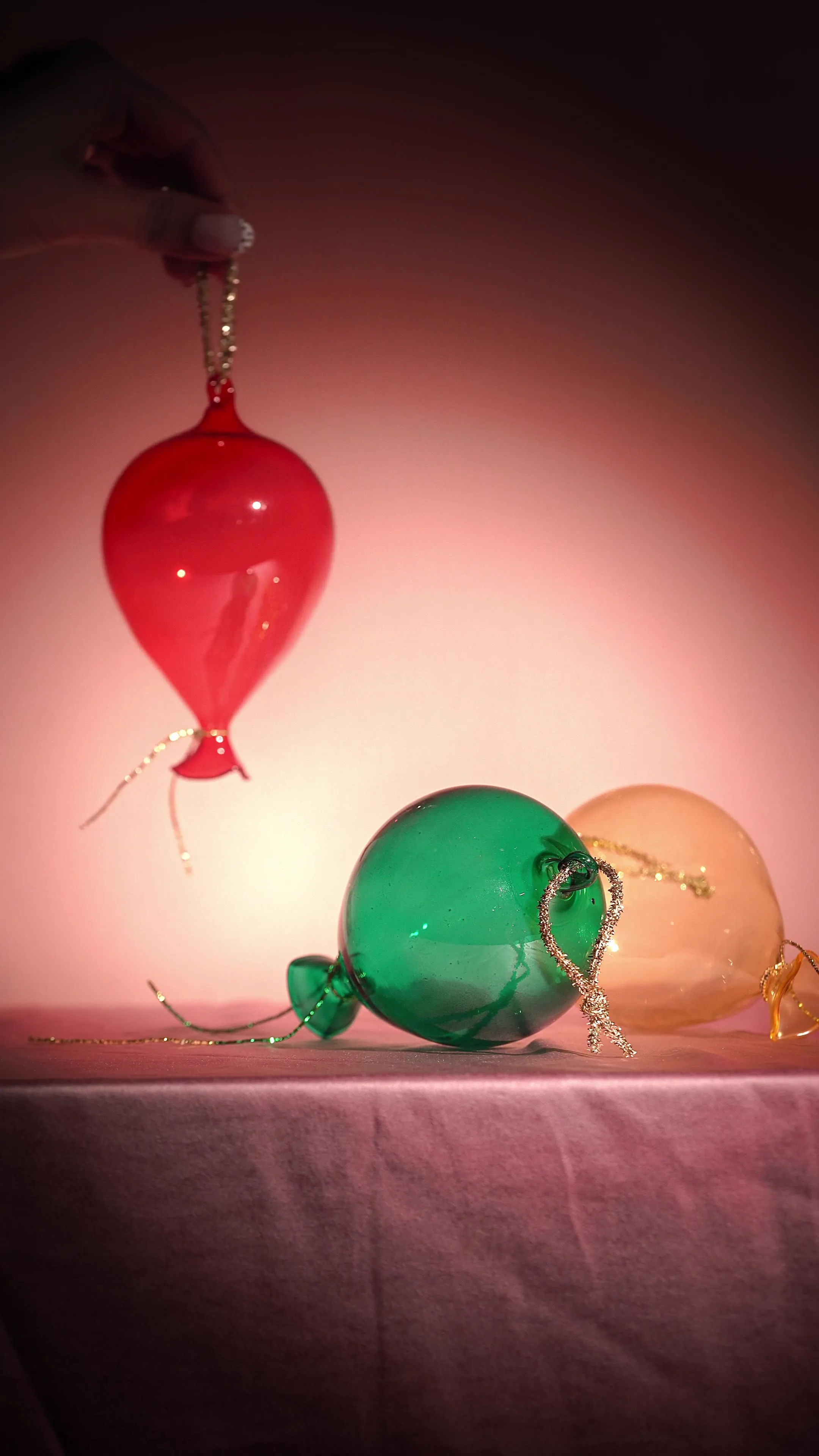 A hand lowering a red glass balloon ornament toward green and amber glass ball ornaments resting on a table.