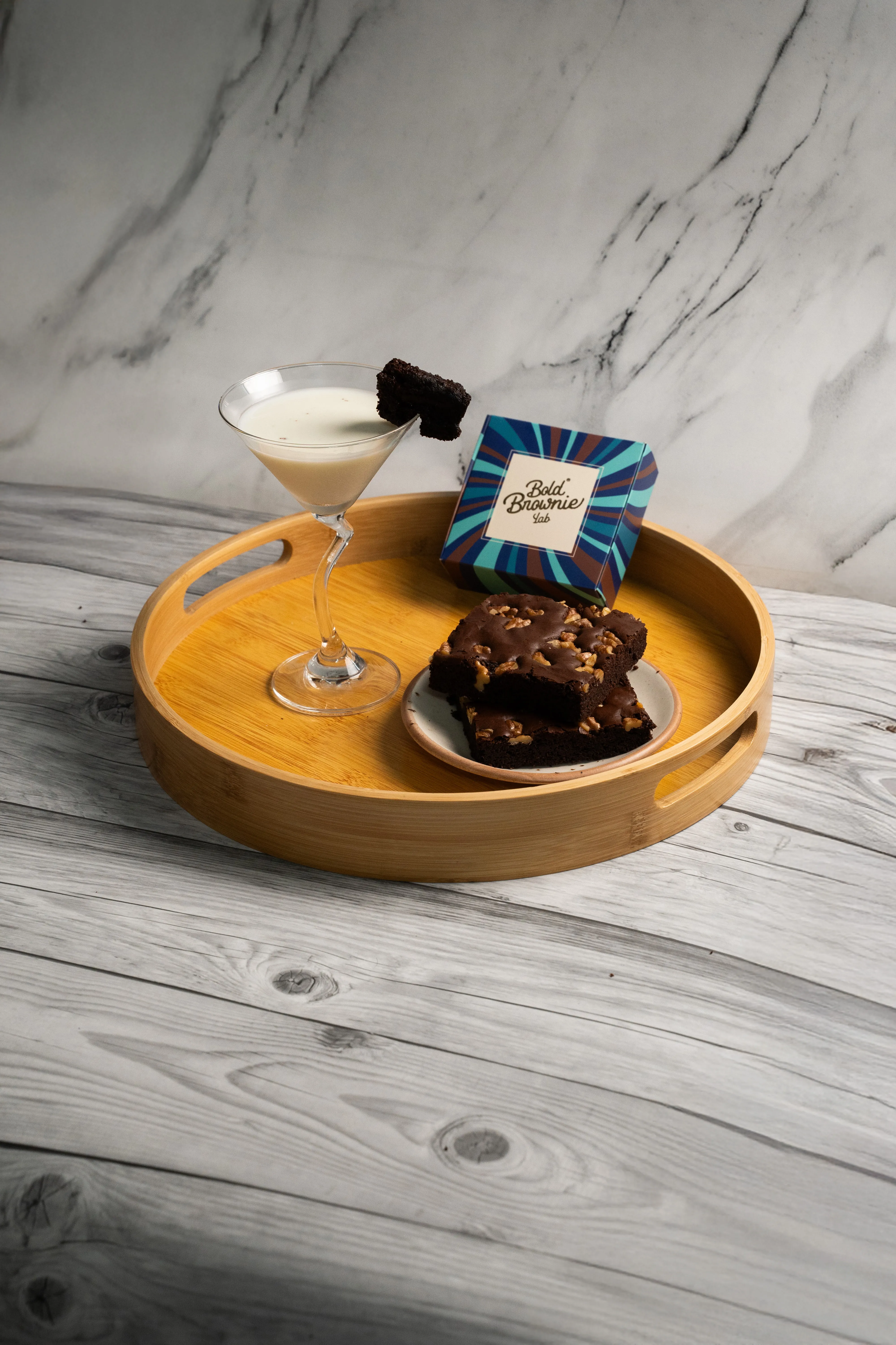 A cream cocktail garnished with a brownie chunk on the rim, beside a Bold Brownie Lab box and a plate of two walnut-topped brownies, on a wooden tray.