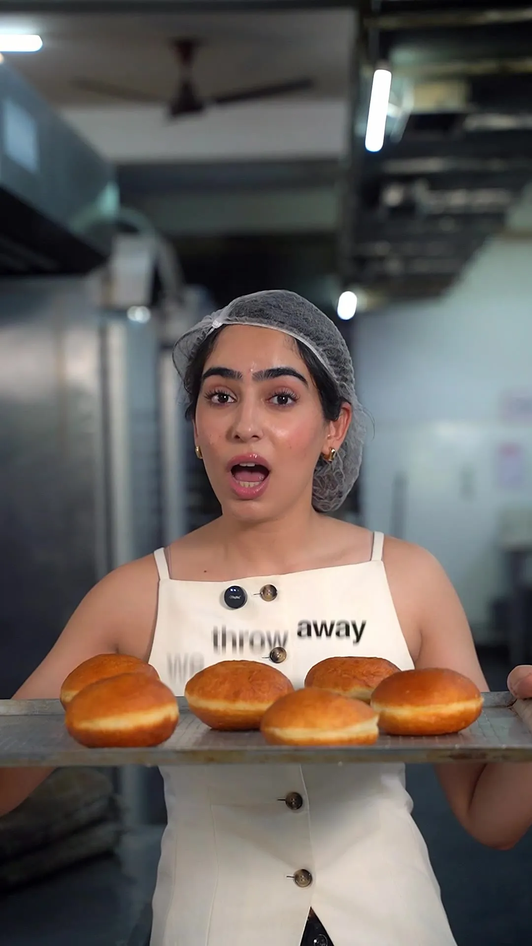 A baker in a hairnet holding a tray of fresh buns, apron captioned "we throw away."