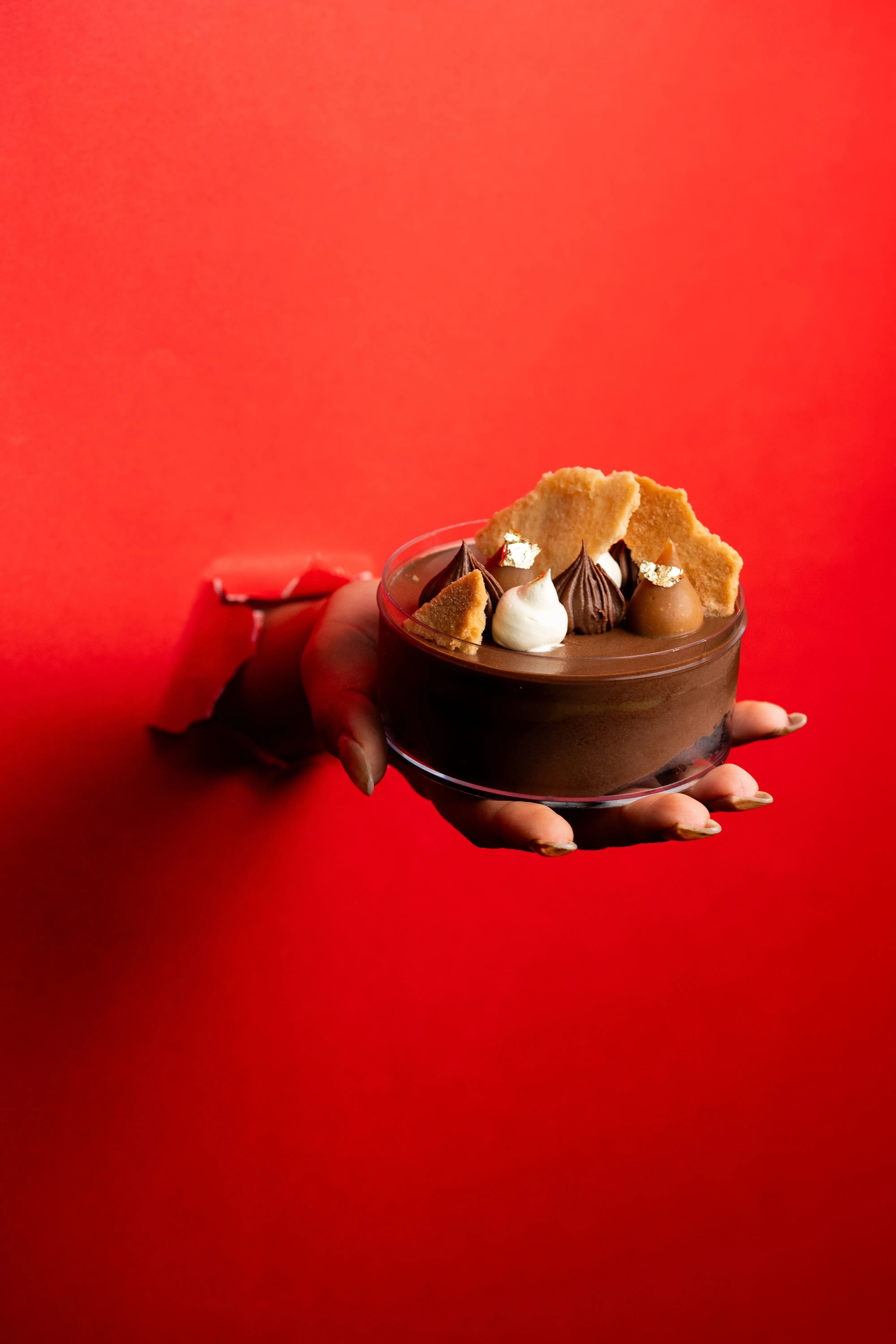 A hand reaching through torn red paper to hold a chocolate mousse dessert topped with shortbread shards, piped cream, and gold leaf.