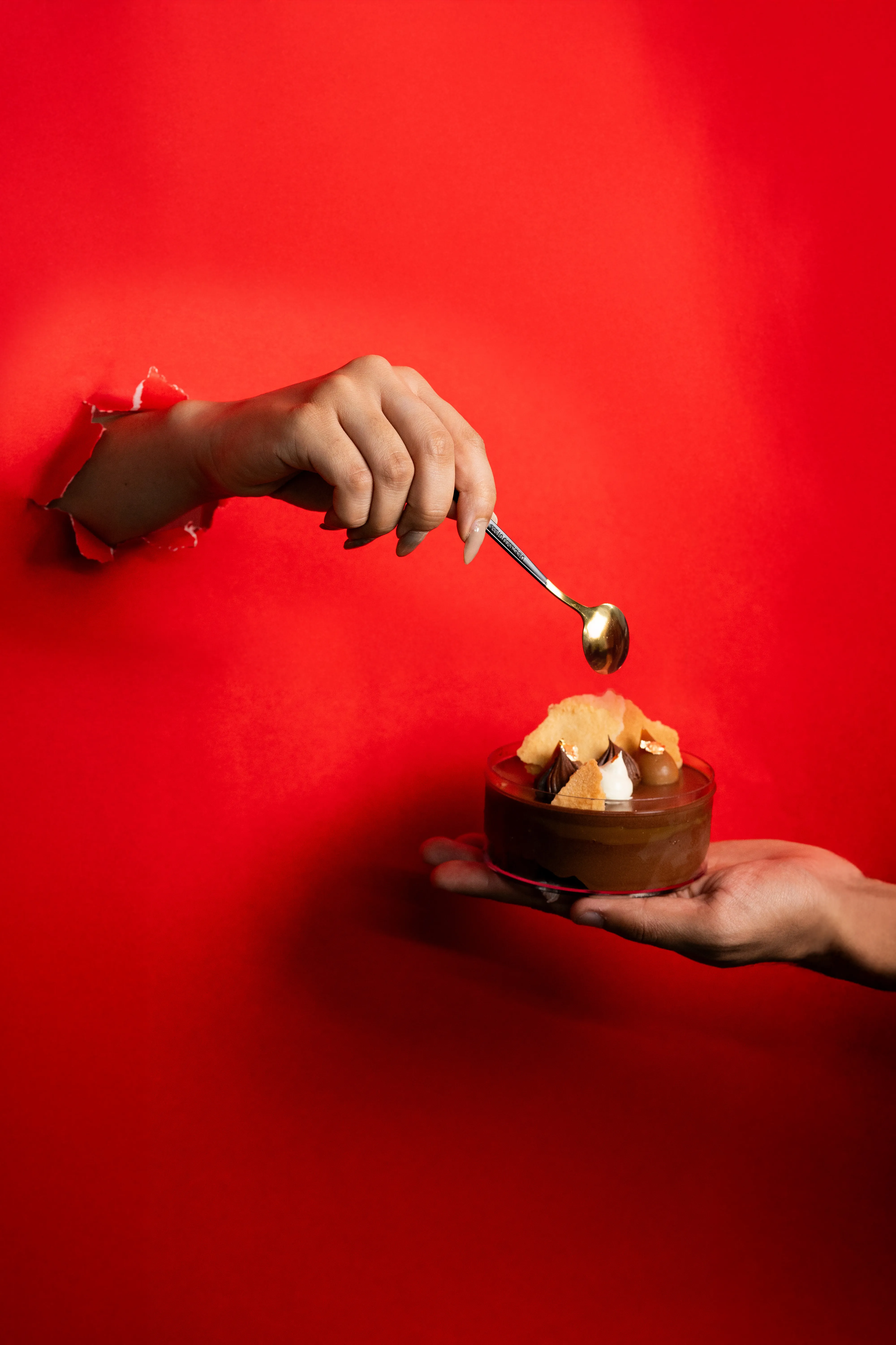 A hand offering the mousse dessert while another hand reaches in with a gold spoon, against a red background.