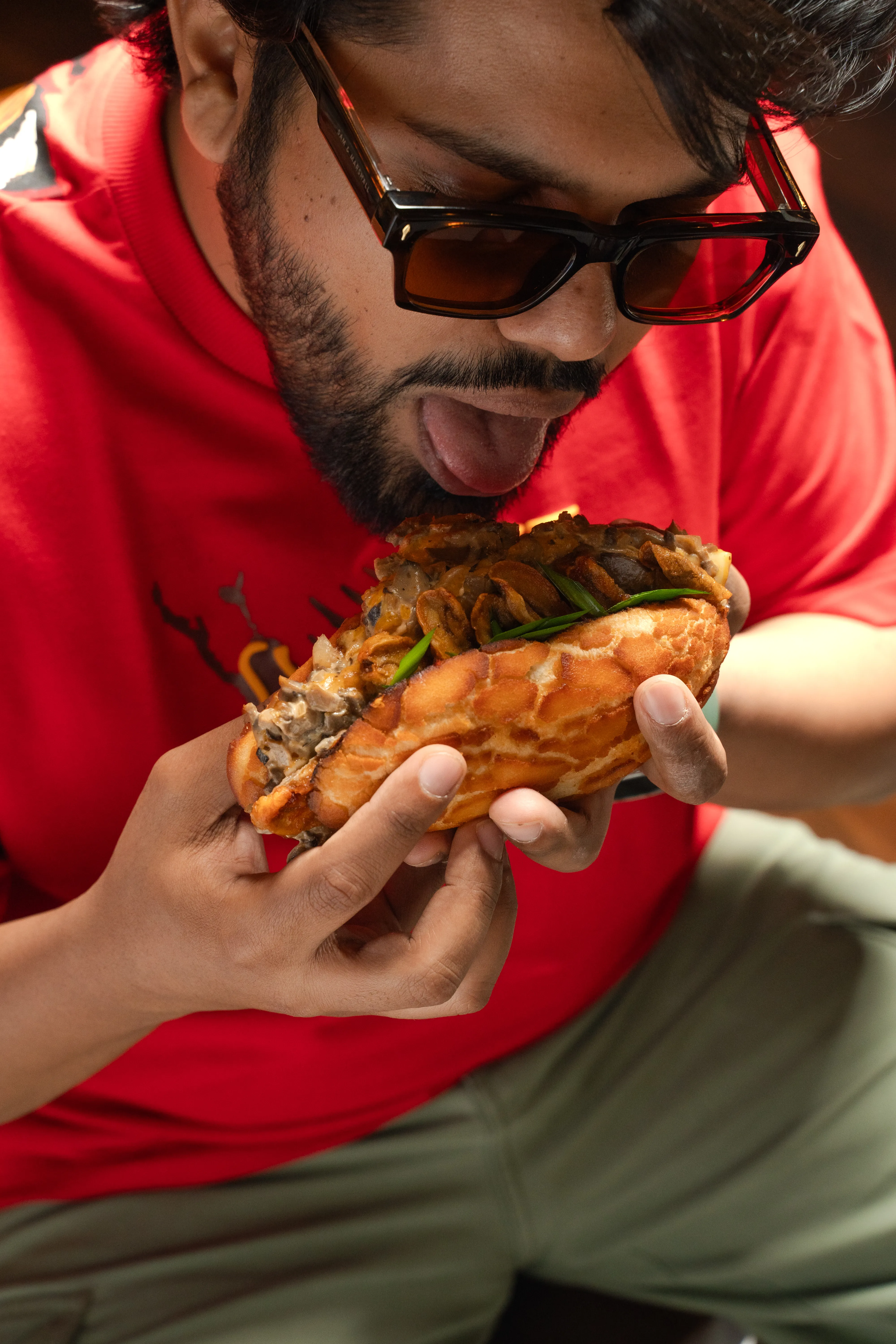 A man in a red t-shirt and sunglasses biting into a mushroom-and-chicken bao sandwich.