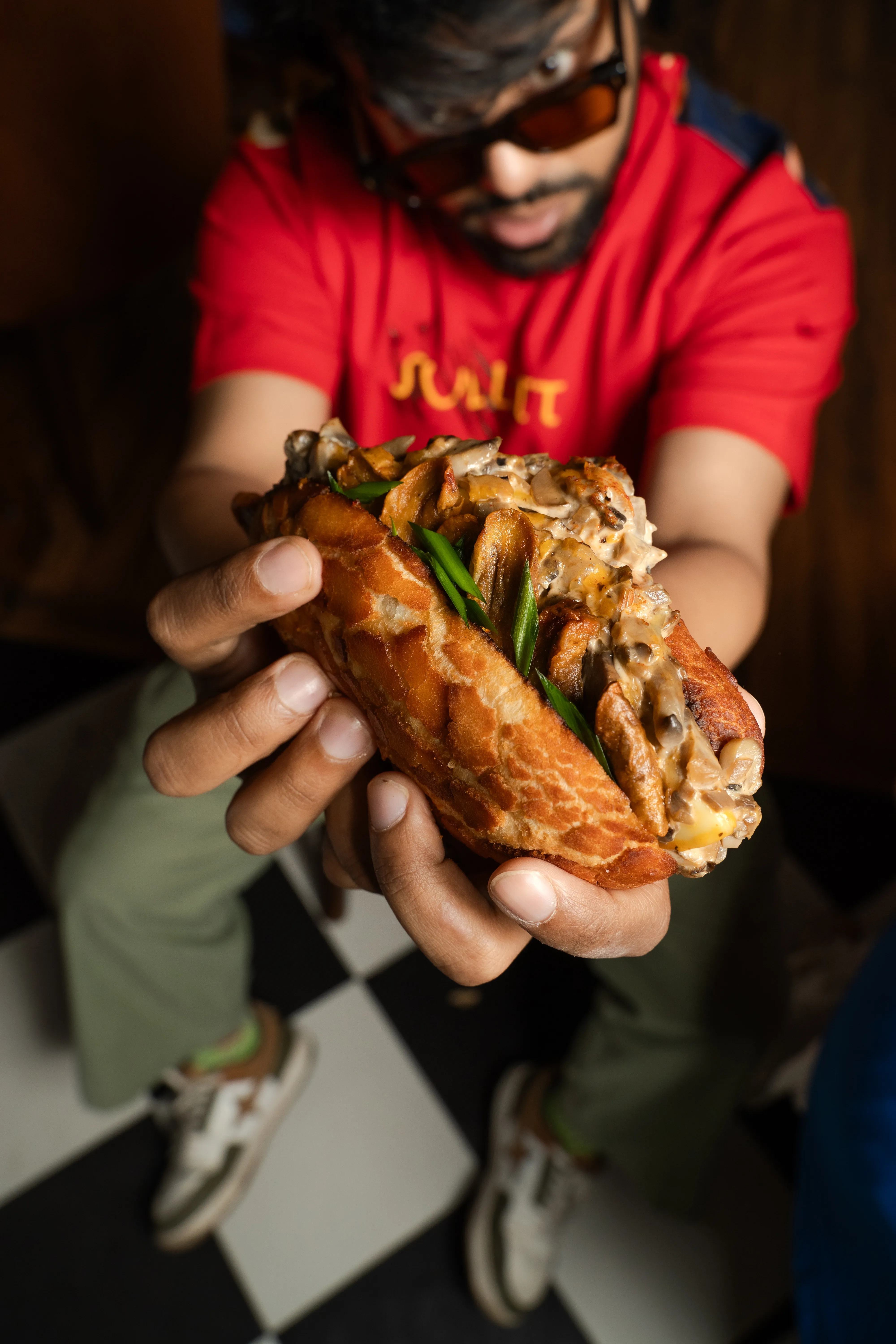 The same man holding the mushroom bao sandwich out toward the camera, seated on a checkerboard floor.