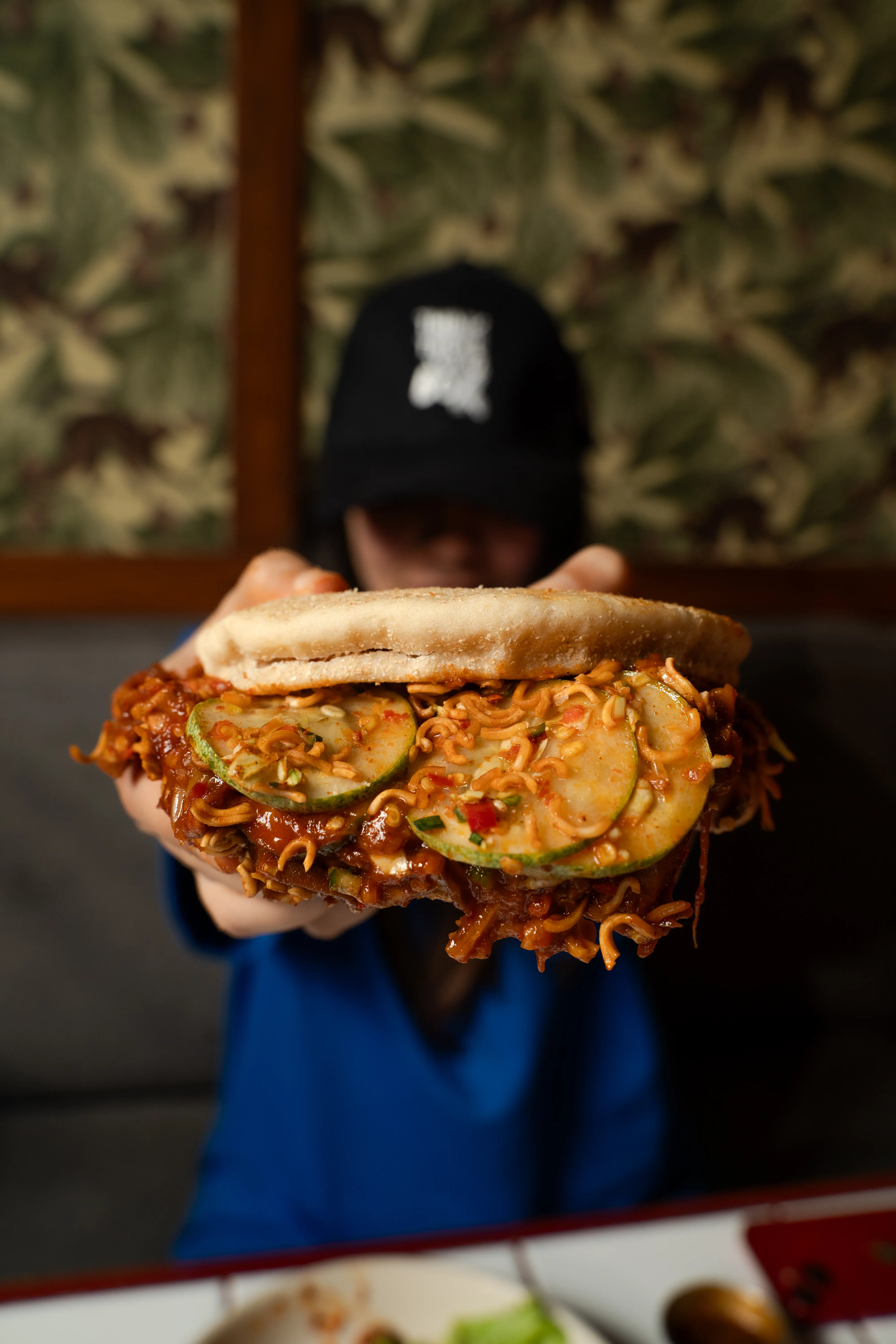 A hand holding out a sloppy joe sandwich piled with ramen noodles and cucumber slices in chili sauce.