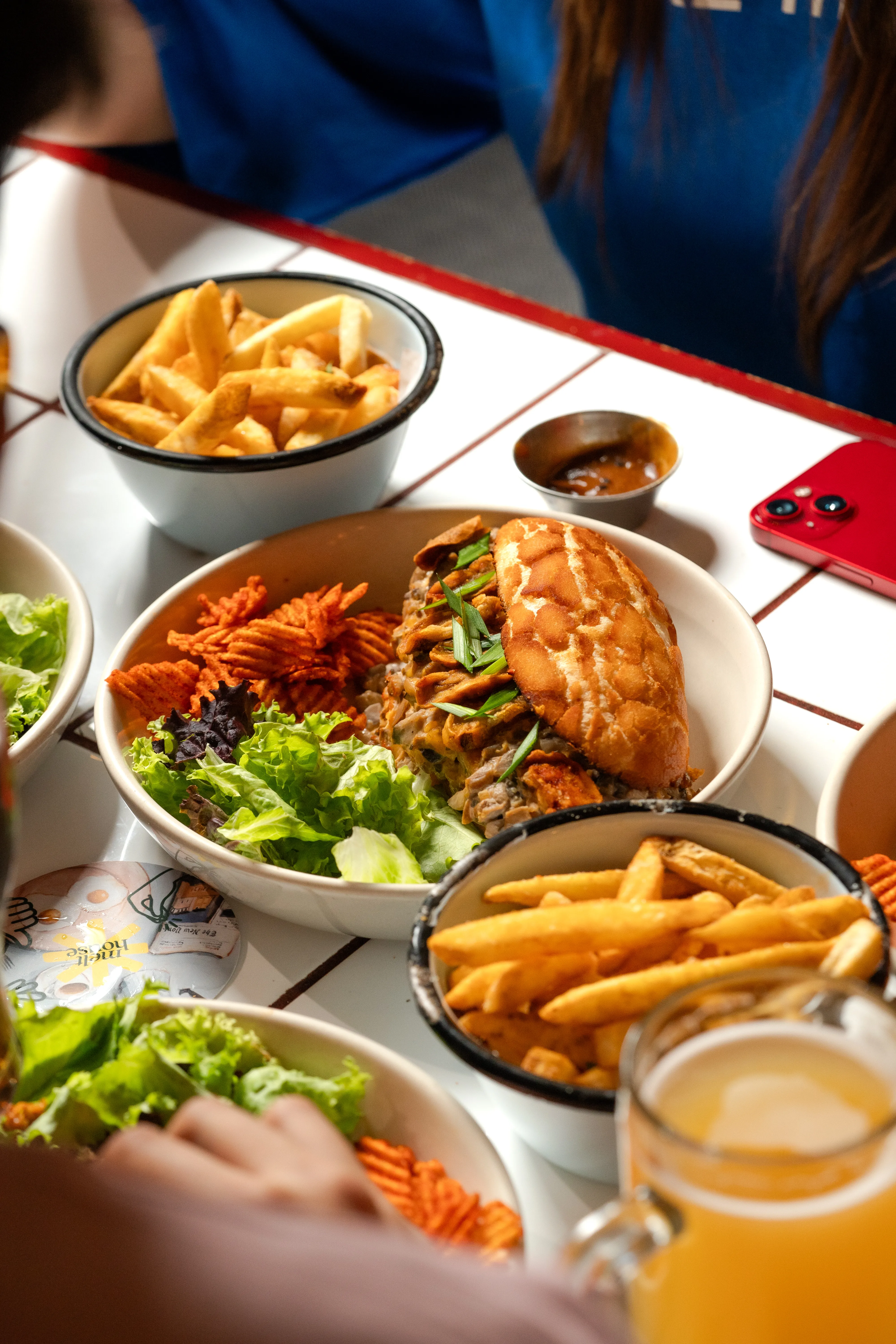 An overhead view of a shared table with the mushroom bao sandwich, a bowl of fries, and a beer mug, a phone resting on the tiles.