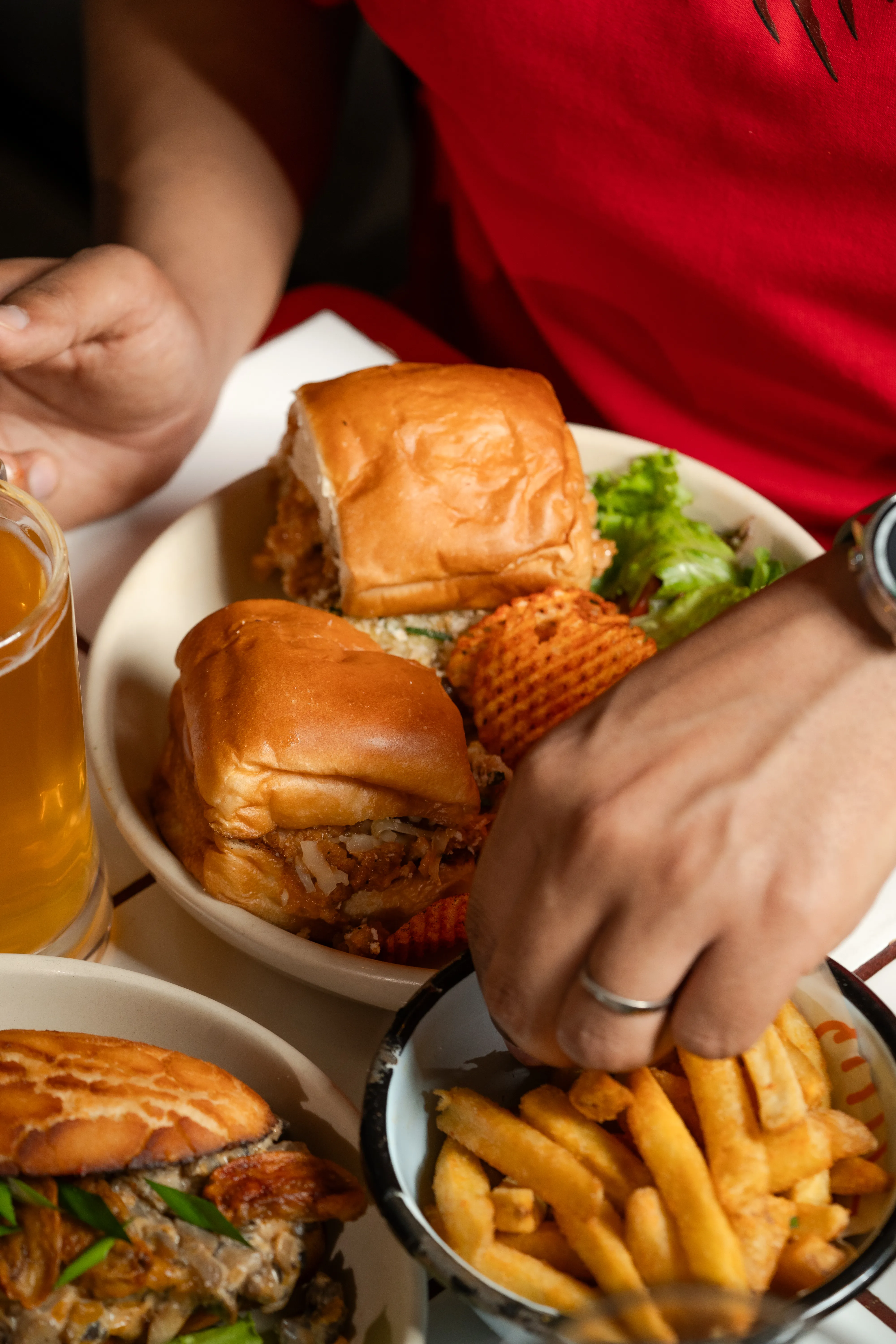 A close-up of hands reaching into a bowl of fries beside a plate of stacked sandwiches, chips, and salad.