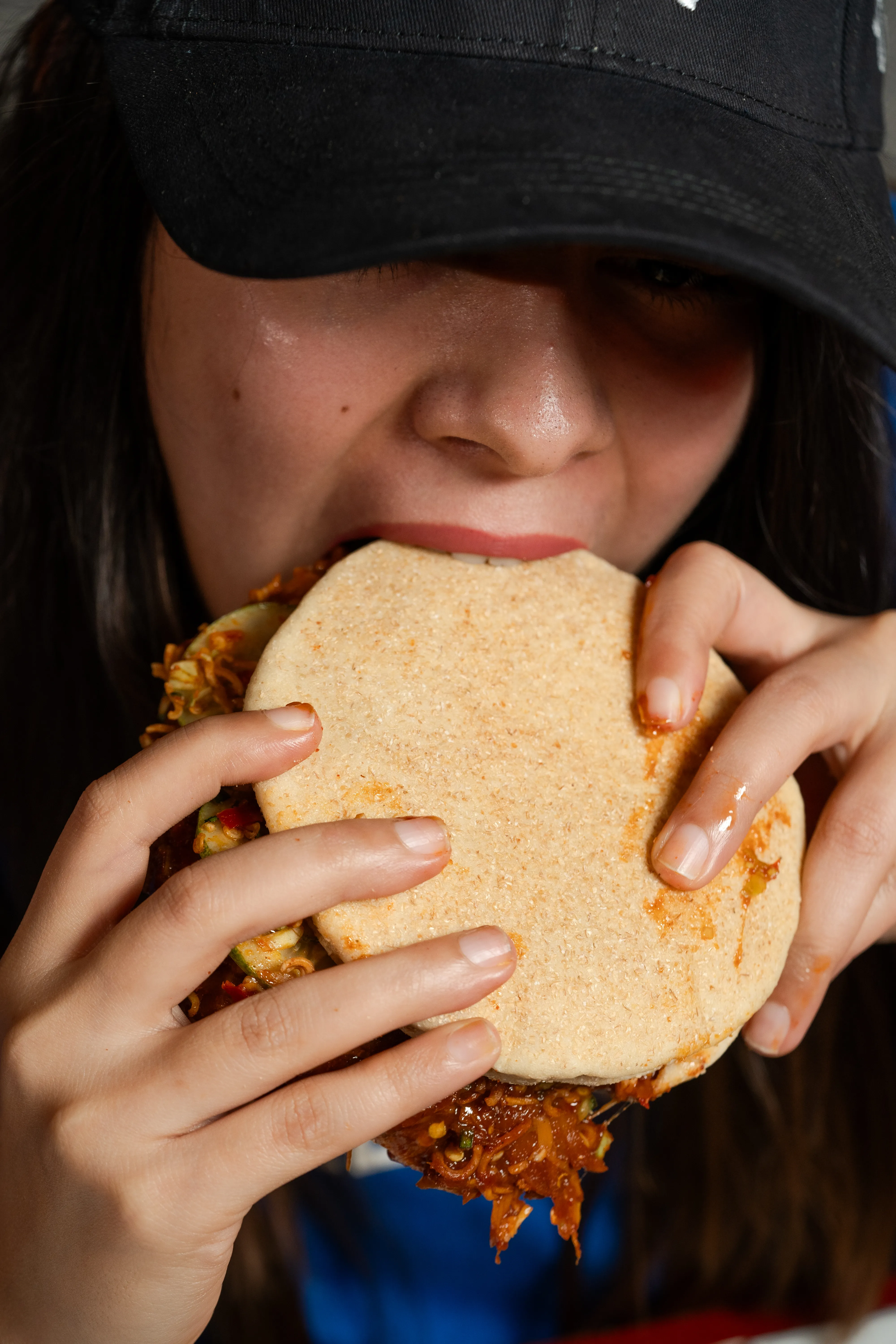 A close-up of a woman in a cap biting into a pita-topped sloppy joe sandwich, ramen and cucumber filling visible.