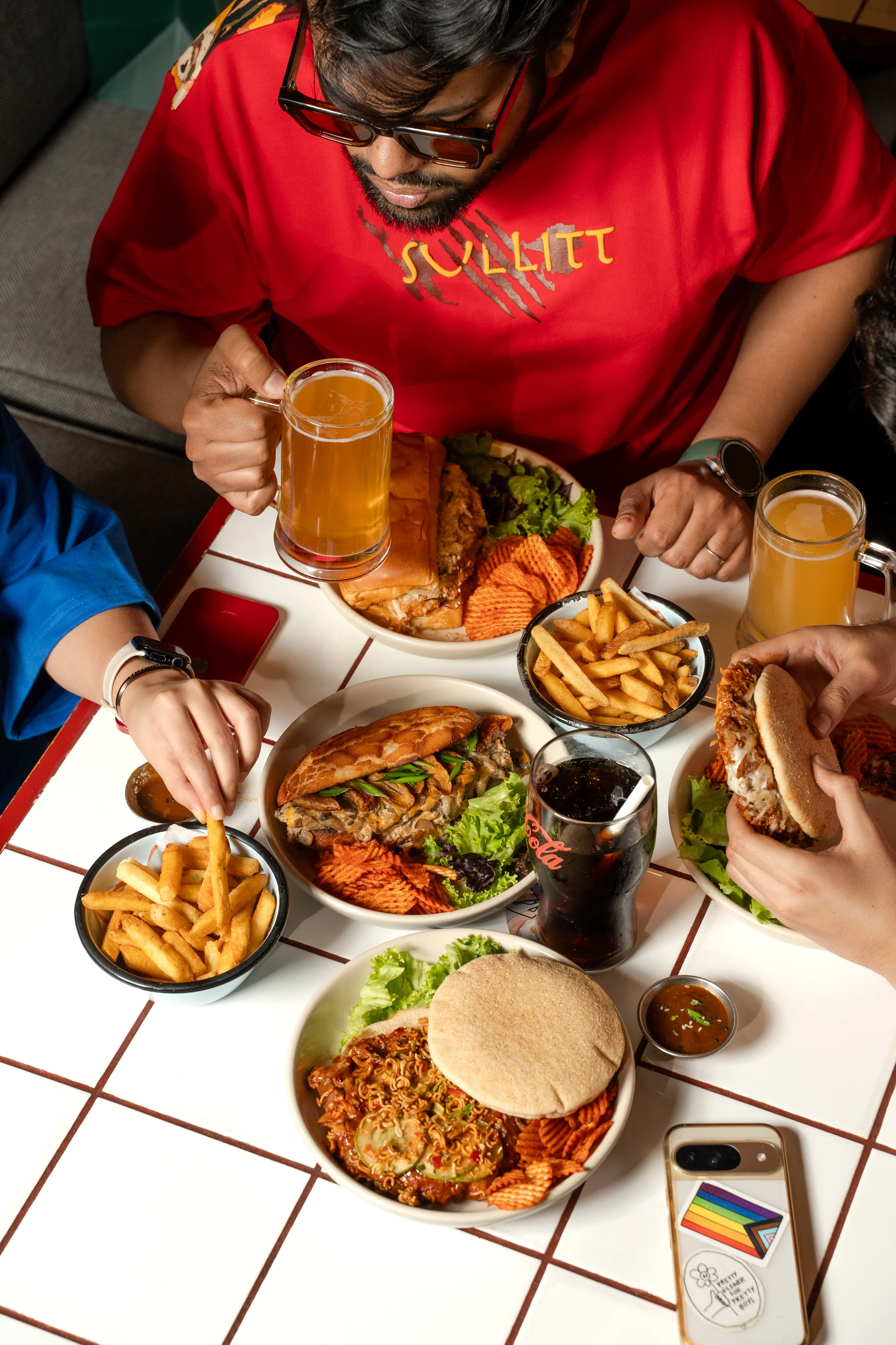 An overhead view of the full table spread — a man raising a beer mug beside bowls of the mushroom bao sandwich, fries, and the ramen sloppy joe sandwich.