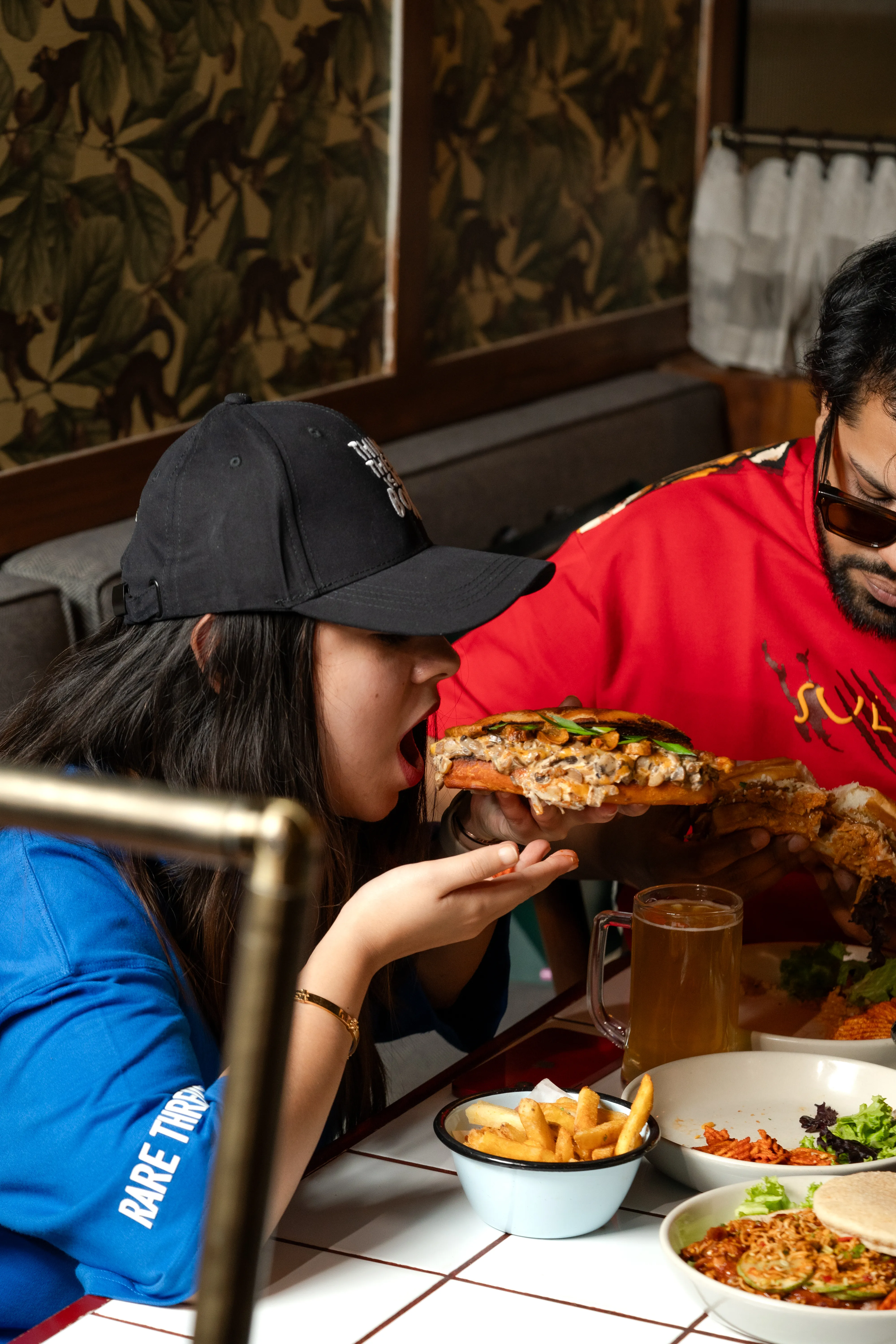 A woman taking a bite of the mushroom bao sandwich held out by a man in sunglasses, beer and fries on the table below.