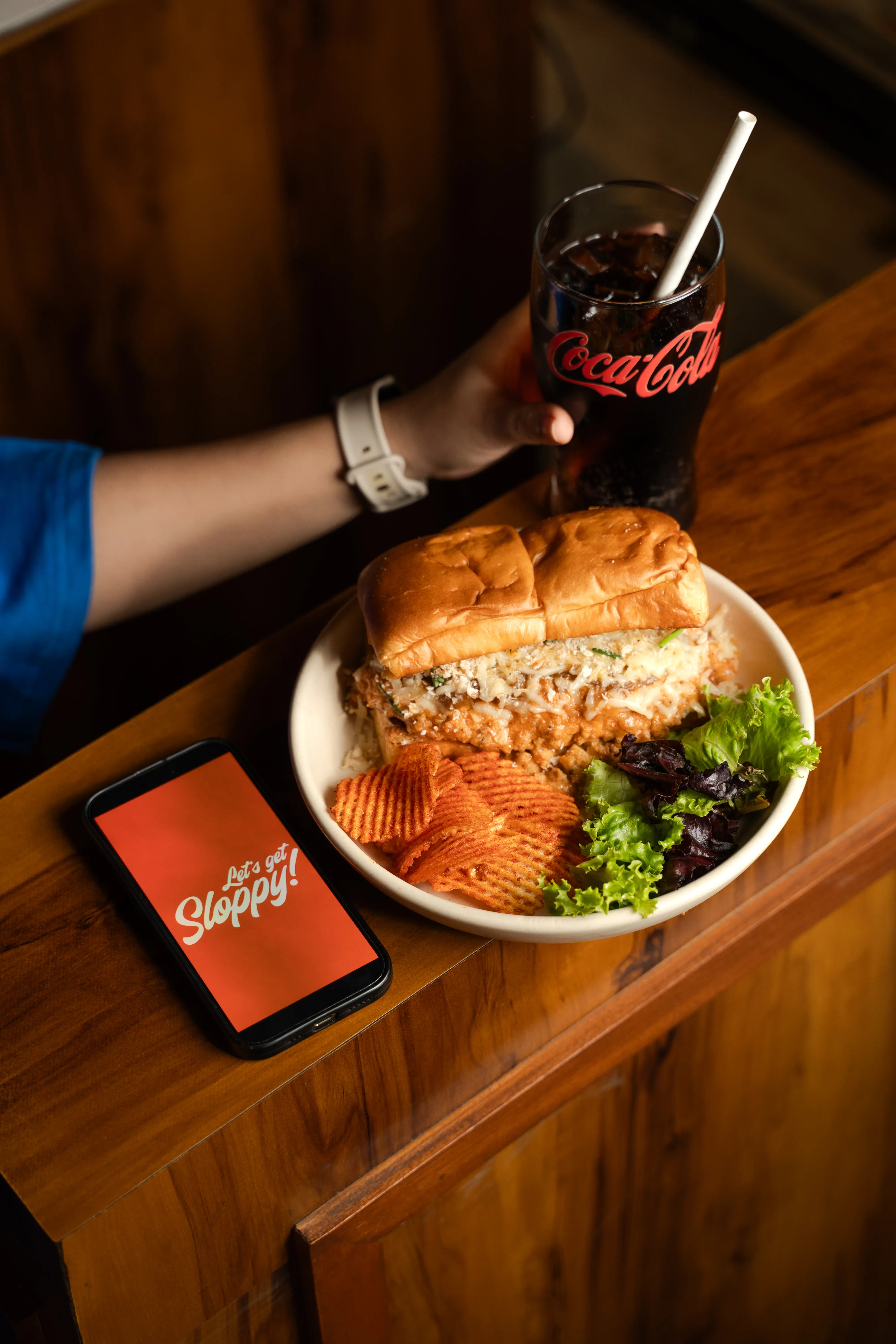 A hand holding a glass of Coca-Cola beside a plated ramen sloppy joe sandwich with chips and salad, a phone reading "Let's get Sloppy!" on the table.