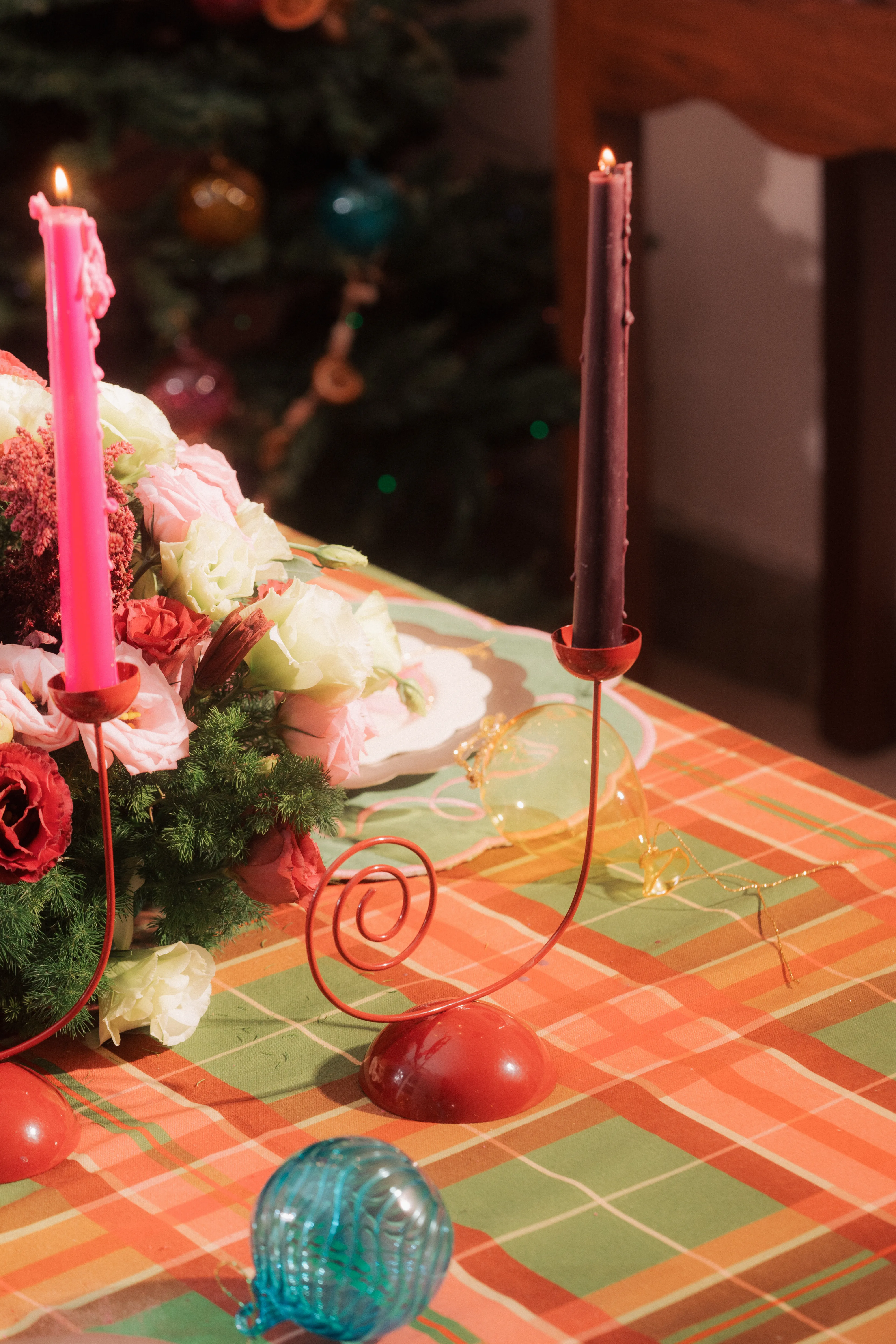 A lit pink and a lit red taper candle, one set in a red spiral candle holder, beside a floral centerpiece on a plaid tablecloth.