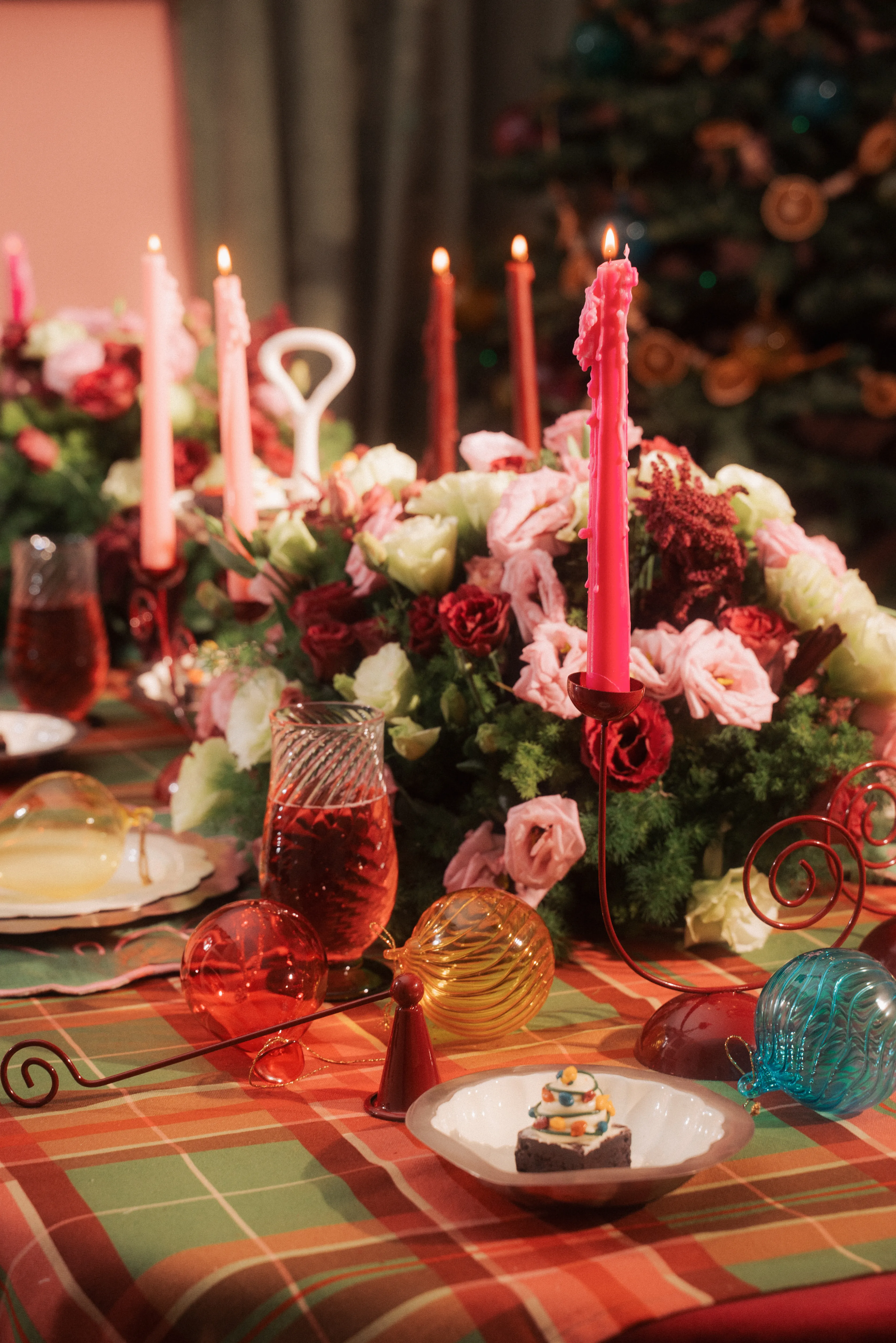 A holiday table spread of lit taper candles, a floral centerpiece, drinks, and glass ball ornaments in front of a Christmas tree.