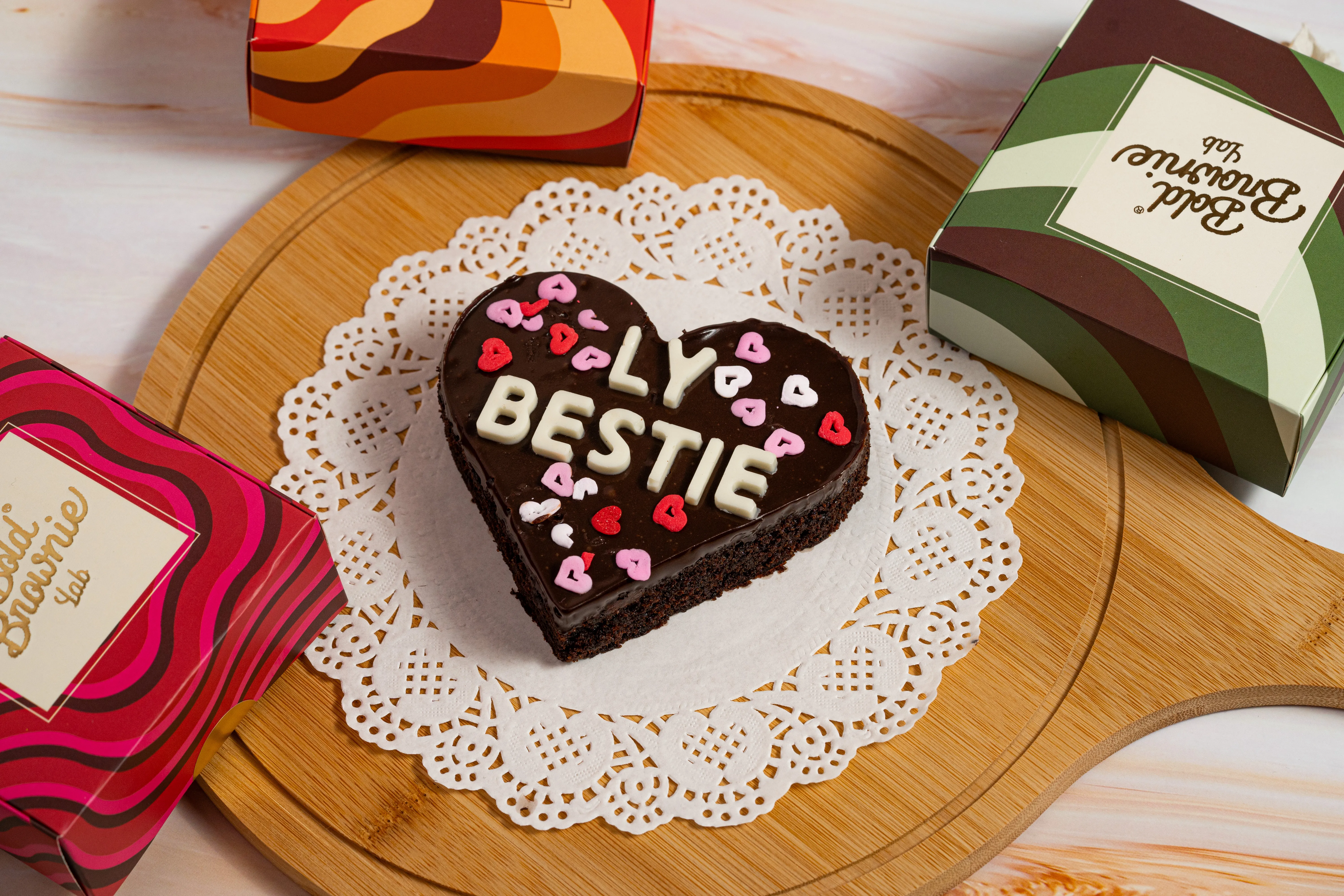 A heart-shaped brownie iced with white chocolate letters reading "BEST BITCH" and rainbow sprinkles, on a lace doily surrounded by Bold Brownie Lab boxes.