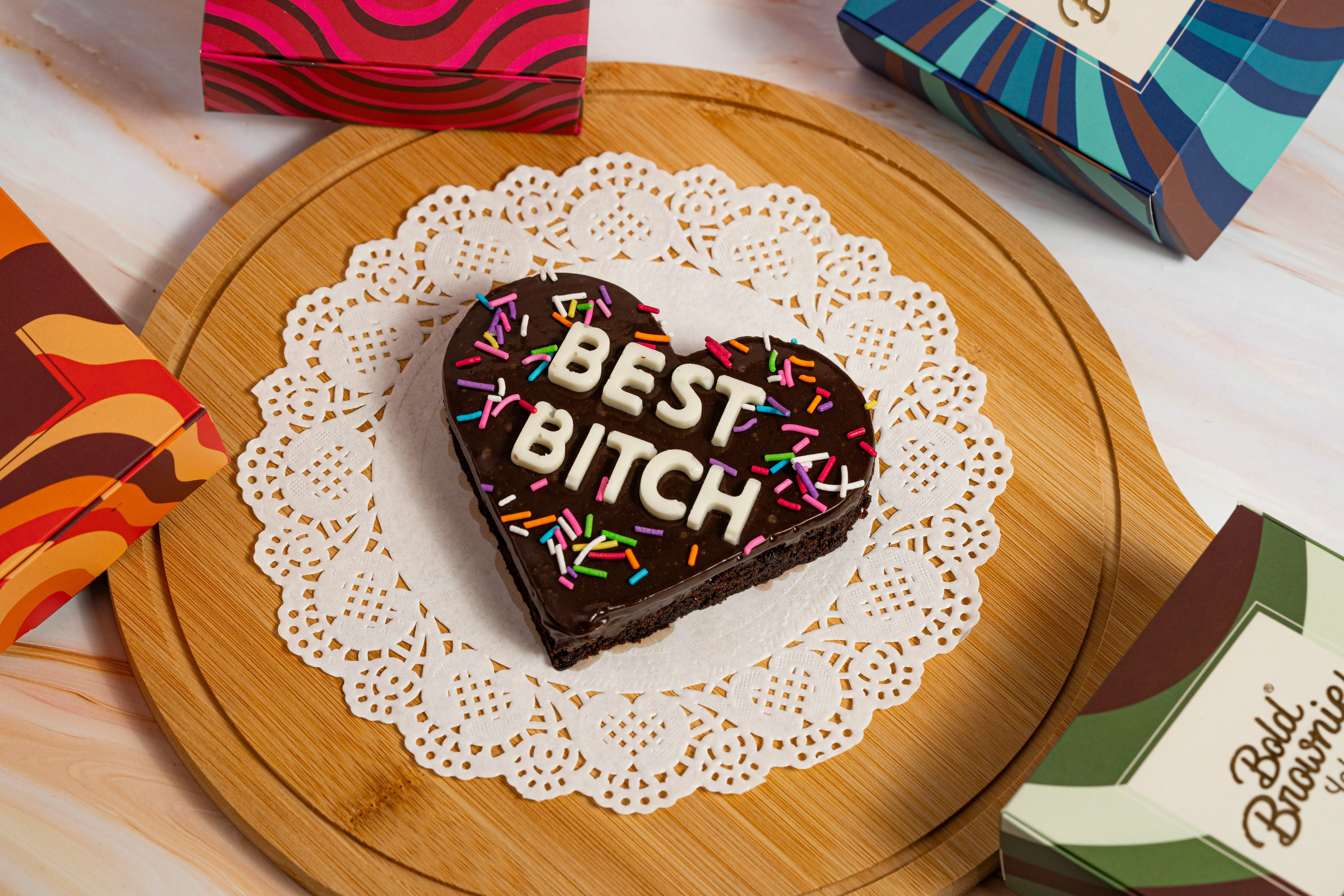 A square brownie iced with white chocolate letters reading "PARTNER IN CRIME" and purple sugar pearls, on a lace doily surrounded by Bold Brownie Lab boxes.