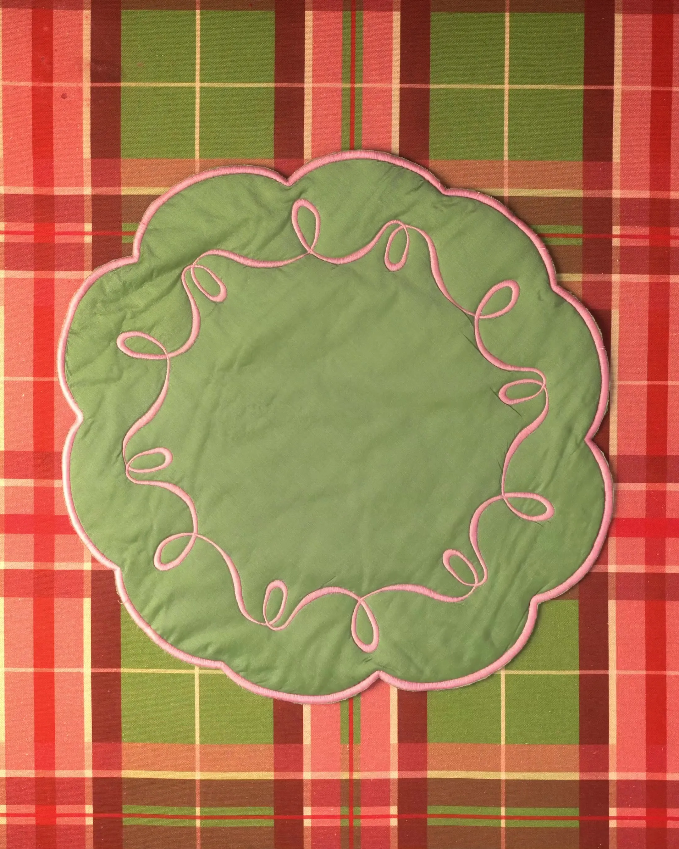 An overhead view of a round pink-and-green quilted placemat with a looped ribbon trim on a plaid tablecloth.