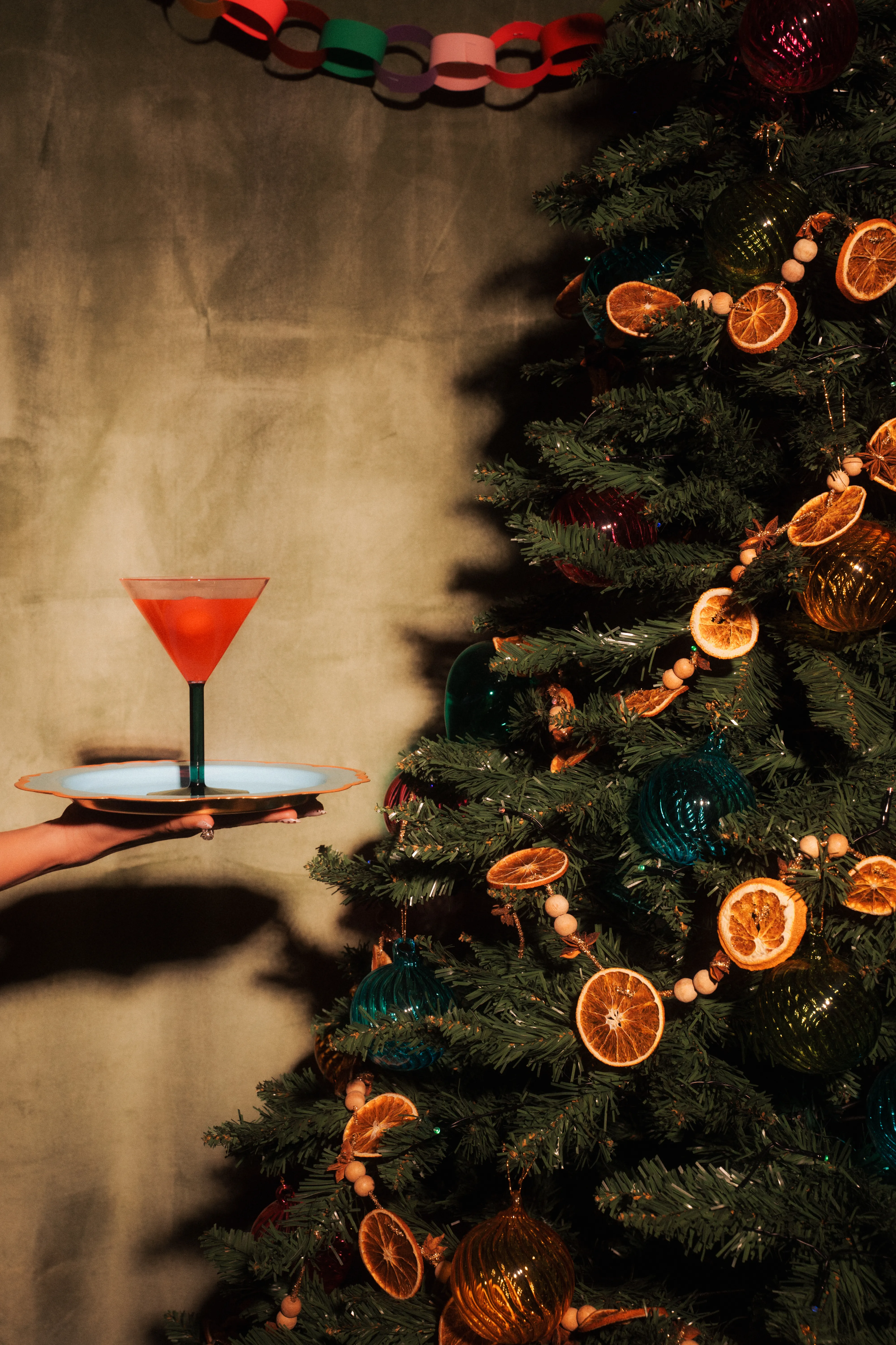 A hand holding out a tray with a red cocktail in front of a Christmas tree strung with dried orange slices and glass ornaments.