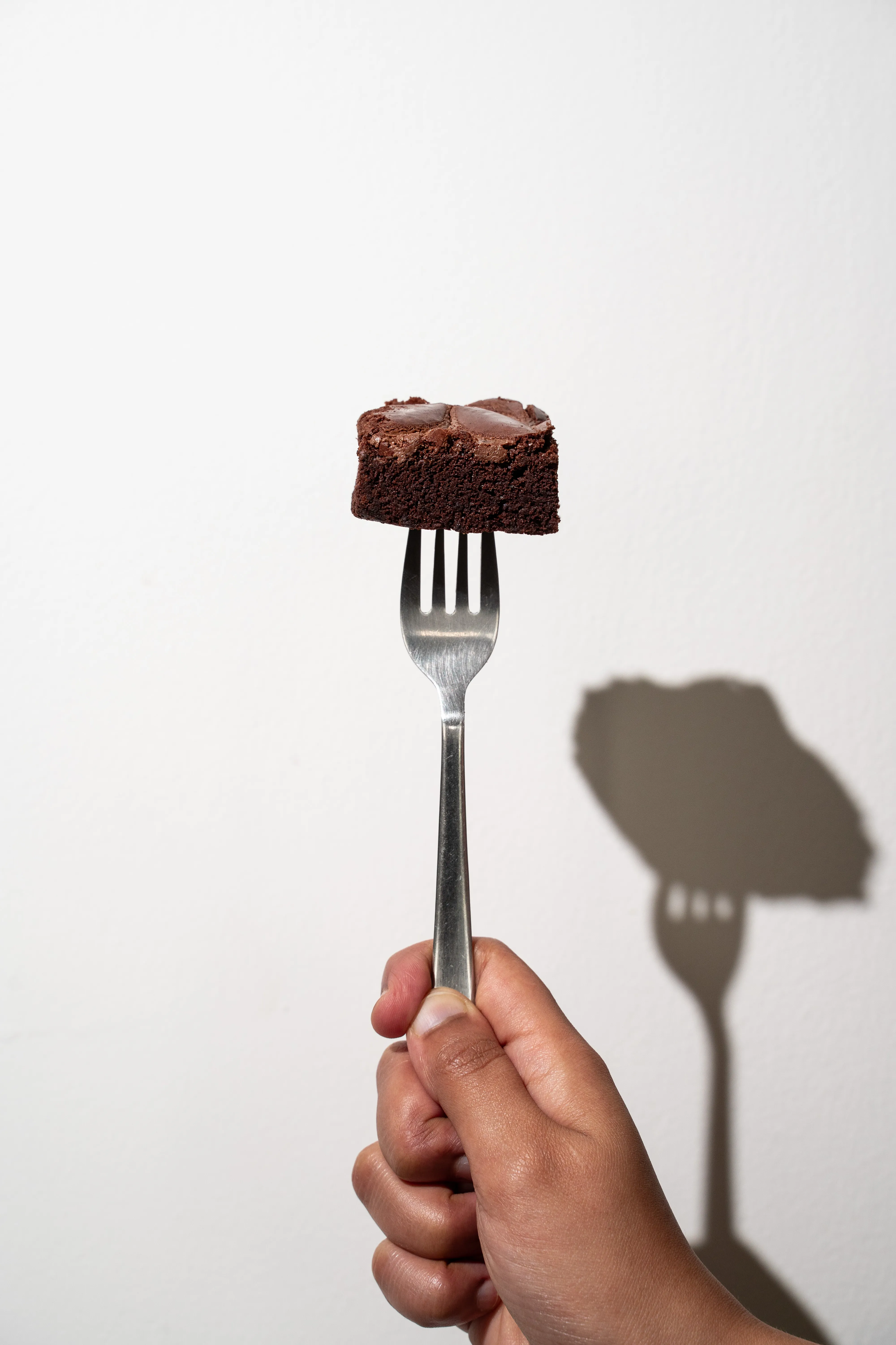 A square of brownie balanced on a fork held up against a plain white wall, casting a long shadow.