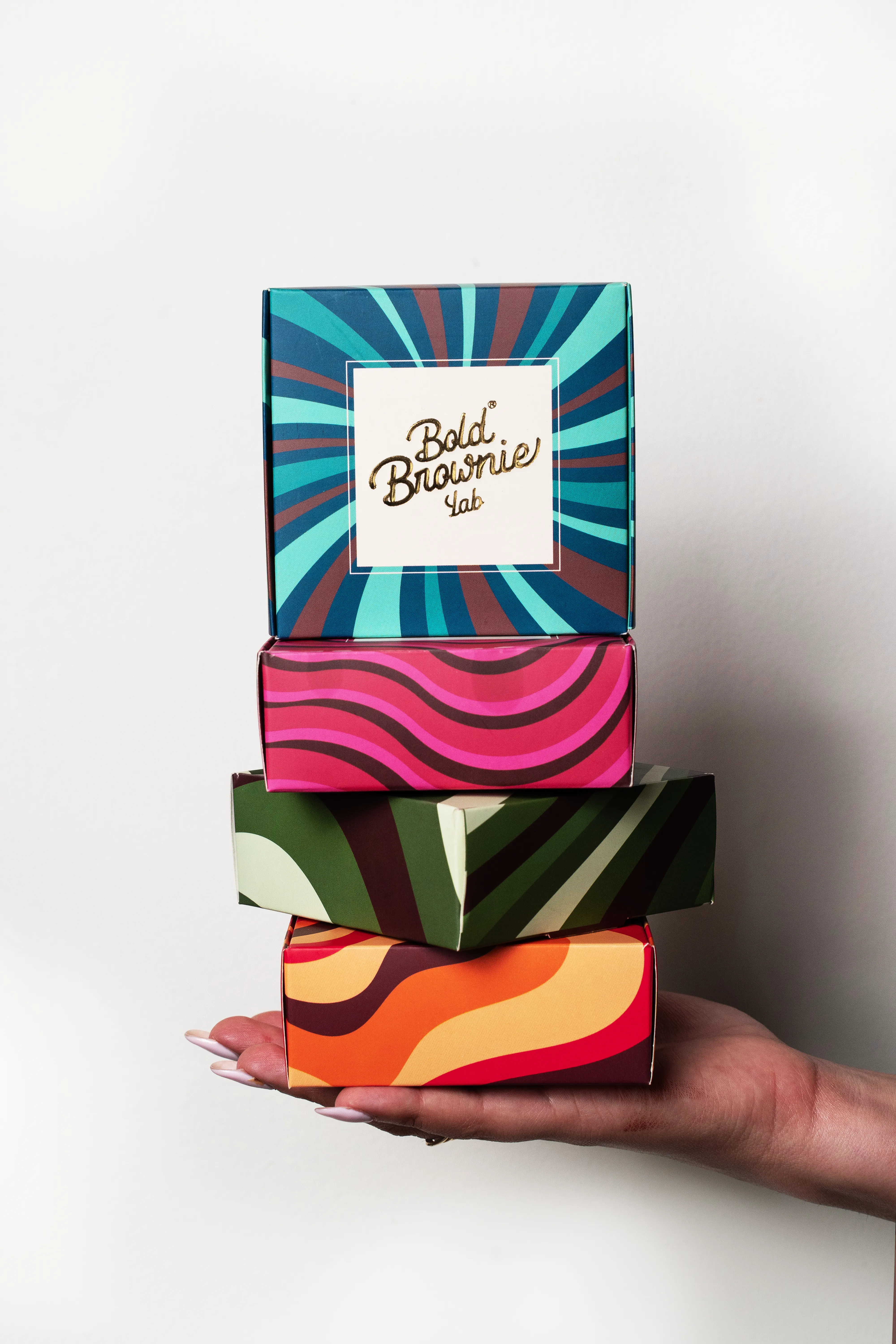 A hand holding a stack of four colorful Bold Brownie Lab boxes against a white background.