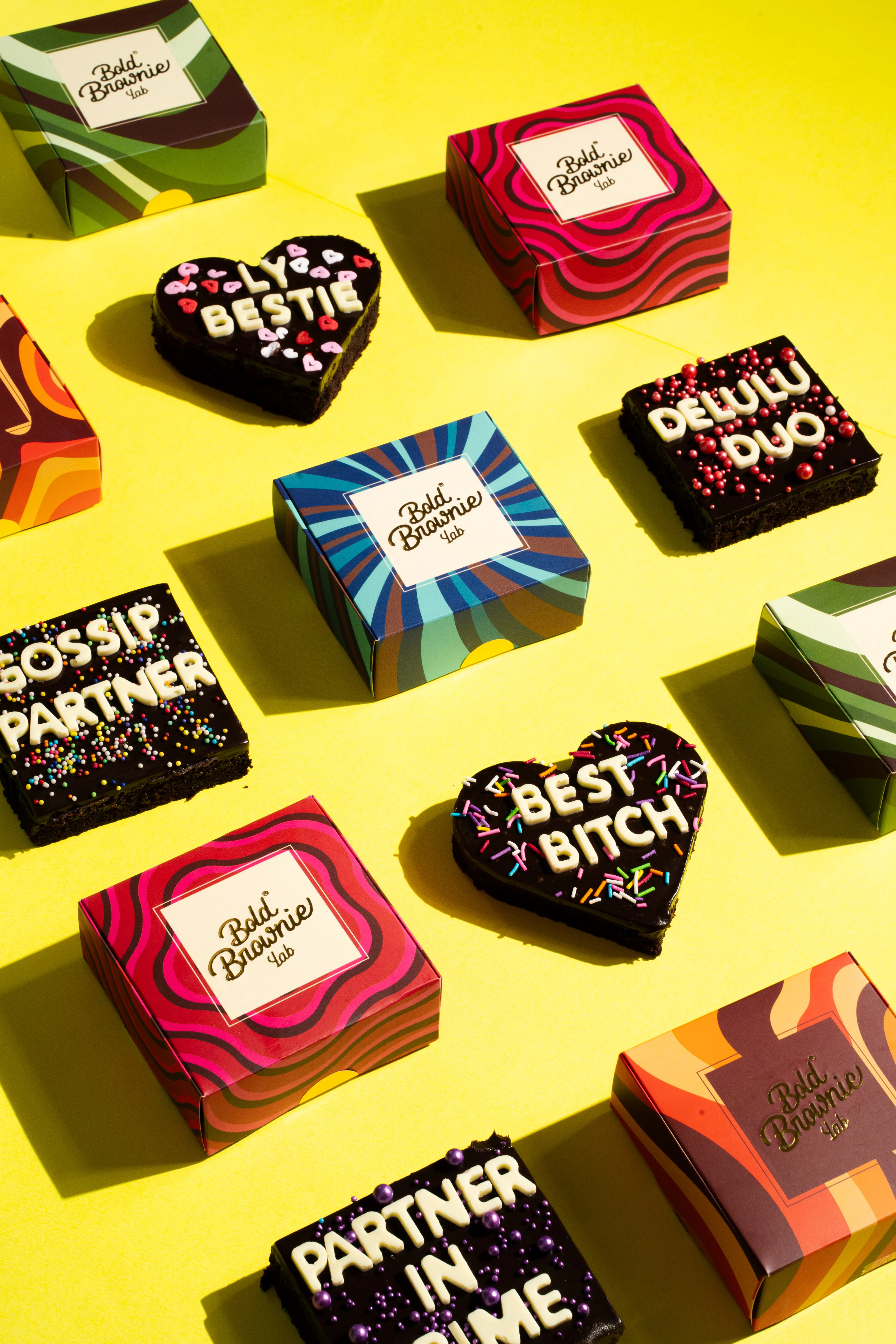 A flat lay of custom text brownies — reading "LY BESTIE," "DELULU DUO," "GOSSIP PARTNER," "BEST BITCH," and "PARTNER IN CRIME" — arranged with Bold Brownie Lab boxes on a yellow background.