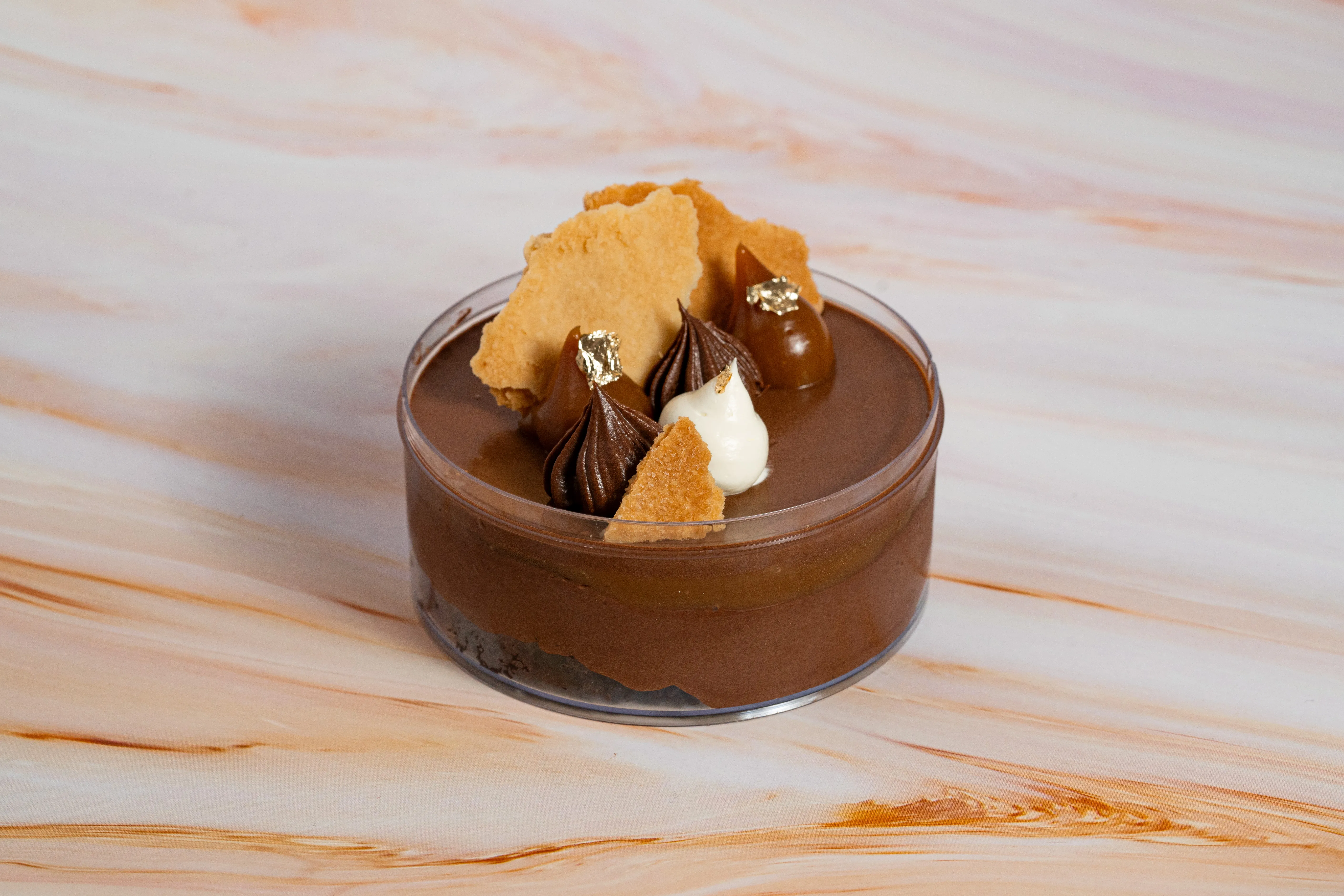 A chocolate mousse dessert in a glass dish, topped with shortbread shards, piped cream and chocolate, and gold leaf, on a marble surface.