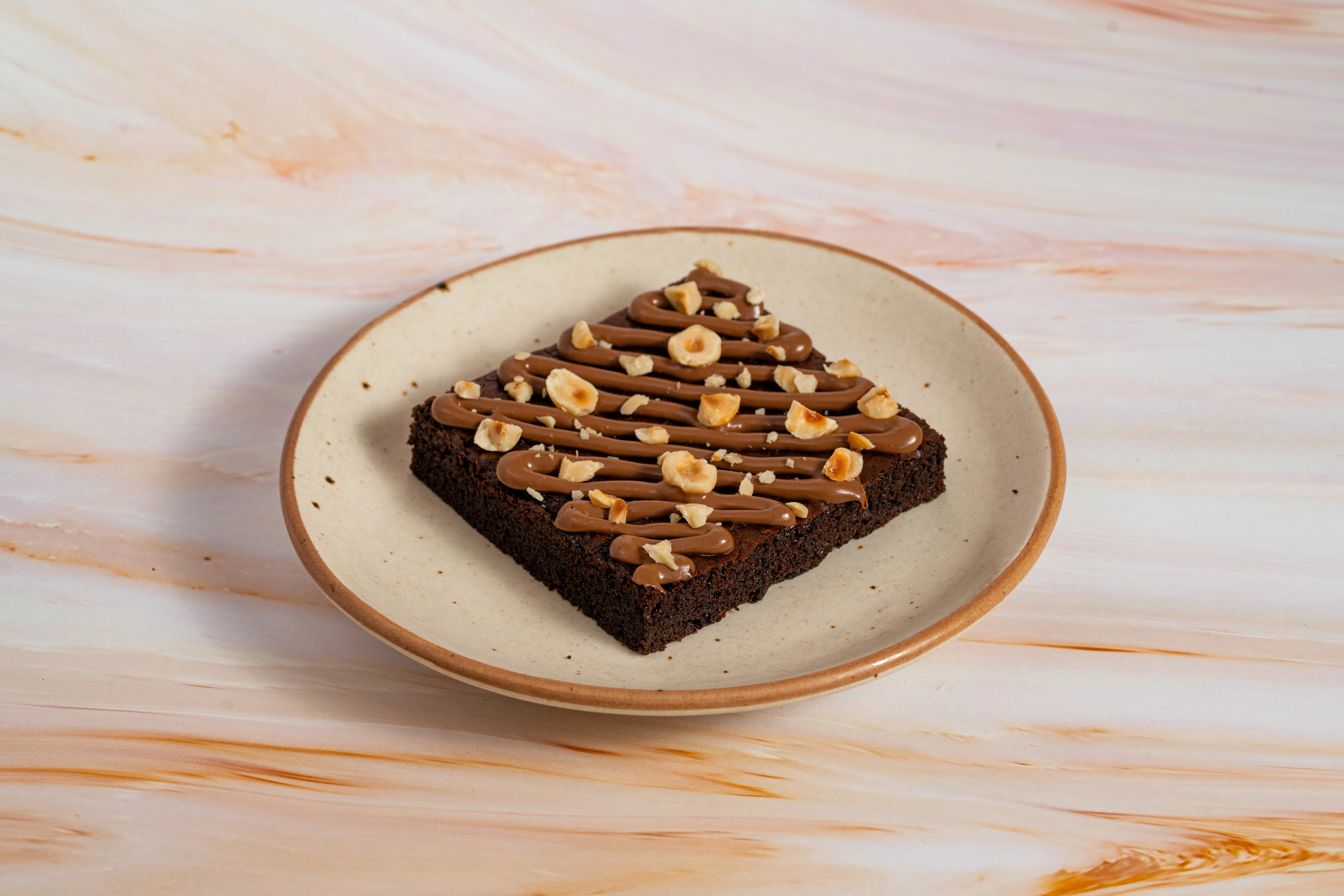 A brownie drizzled with chocolate and topped with hazelnuts, plated on a marble surface, from another angle.